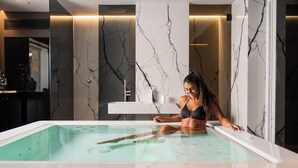 Una mujer con traje de baño negro sentada en una bañera moderna con revestimiento de mármol y cerramiento de vidrio.