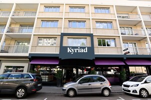 Un hotel de varias plantas llamado 'Kyriad', con una fachada moderna, balcones y un estacionamiento con varios autos.