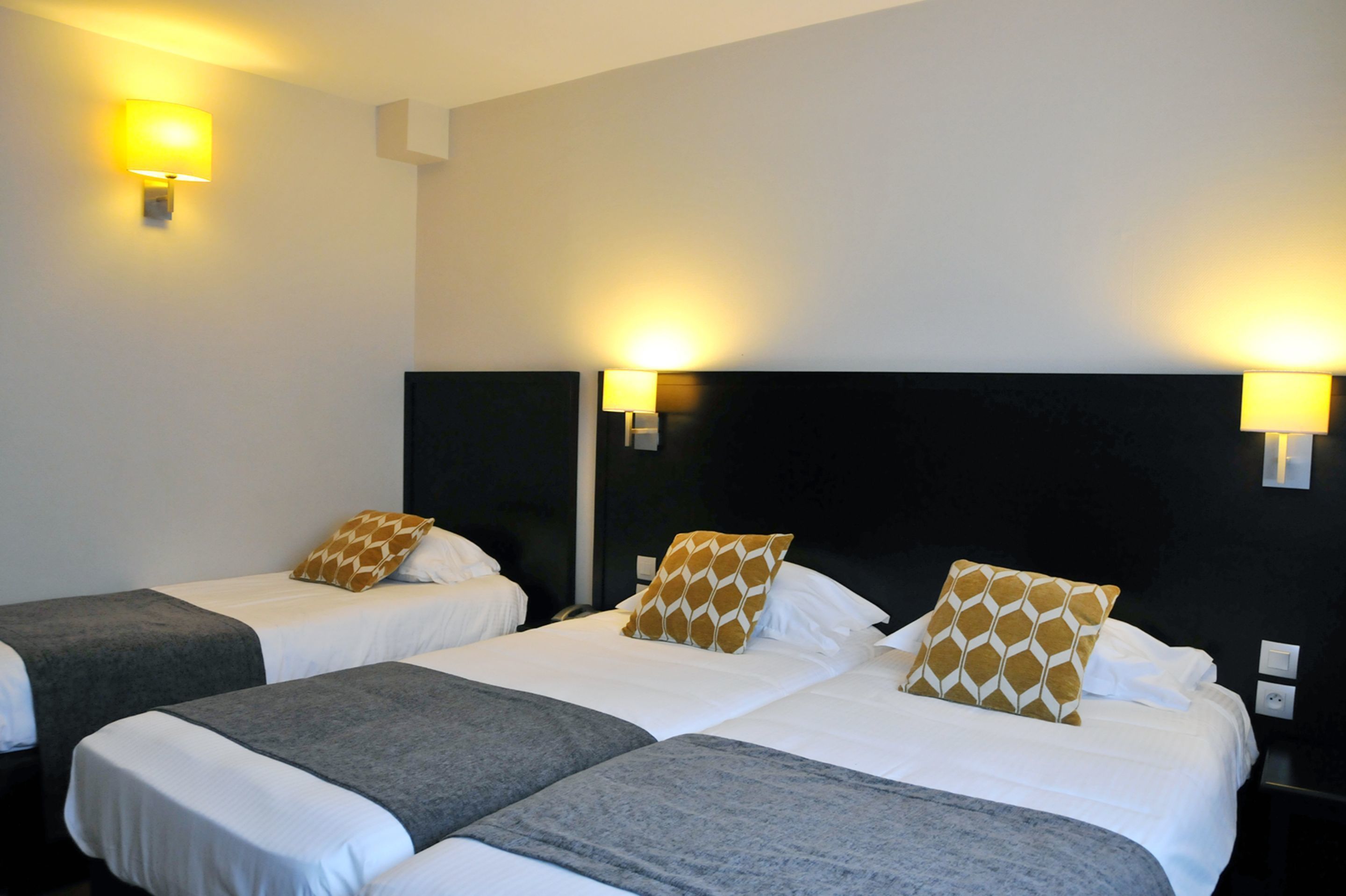 A hotel room with three single beds, each with a grey blanket and a patterned pillow. There are two wall-mounted lamps providing light.