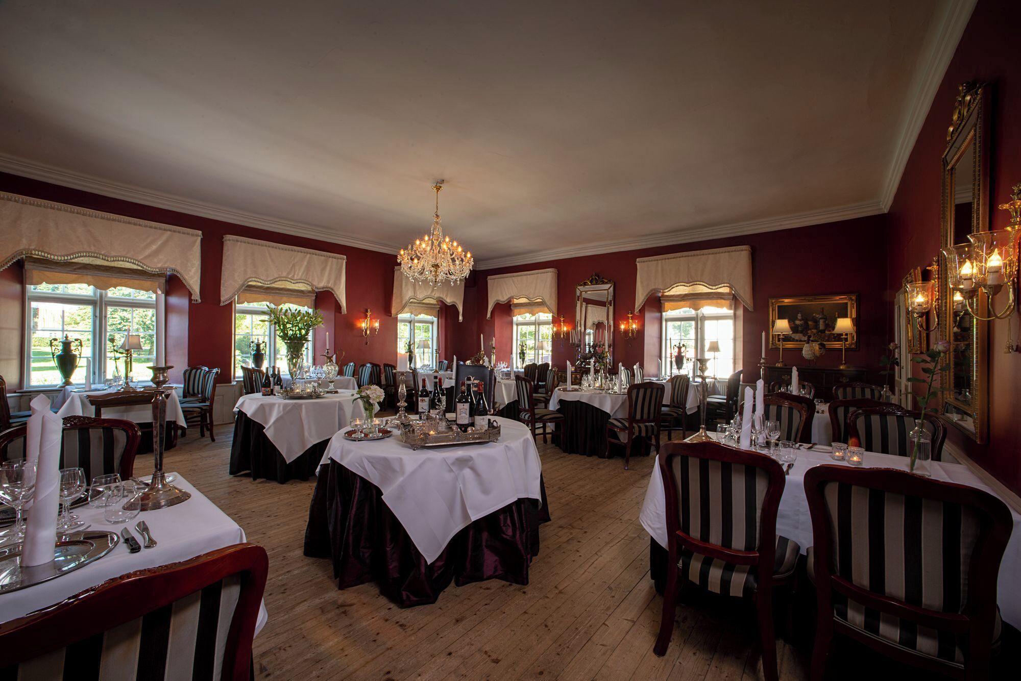 A formal dining room with multiple tables set for service, large windows with curtains, and a chandelier.