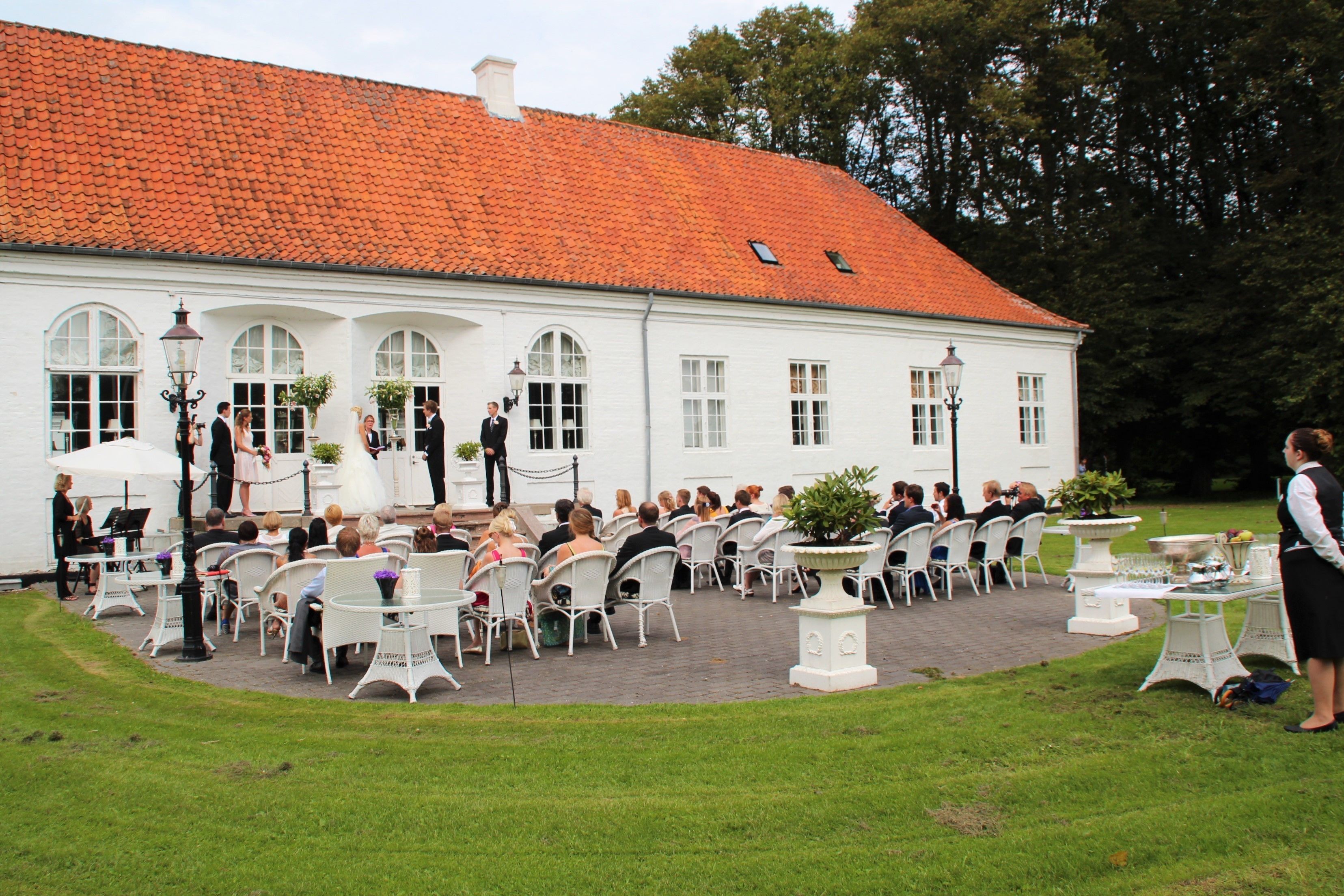 Een wit gebouw met een rode pannendak, een buiten zitgedeelte met witte stoelen en tafels, en een geplaveid pad.