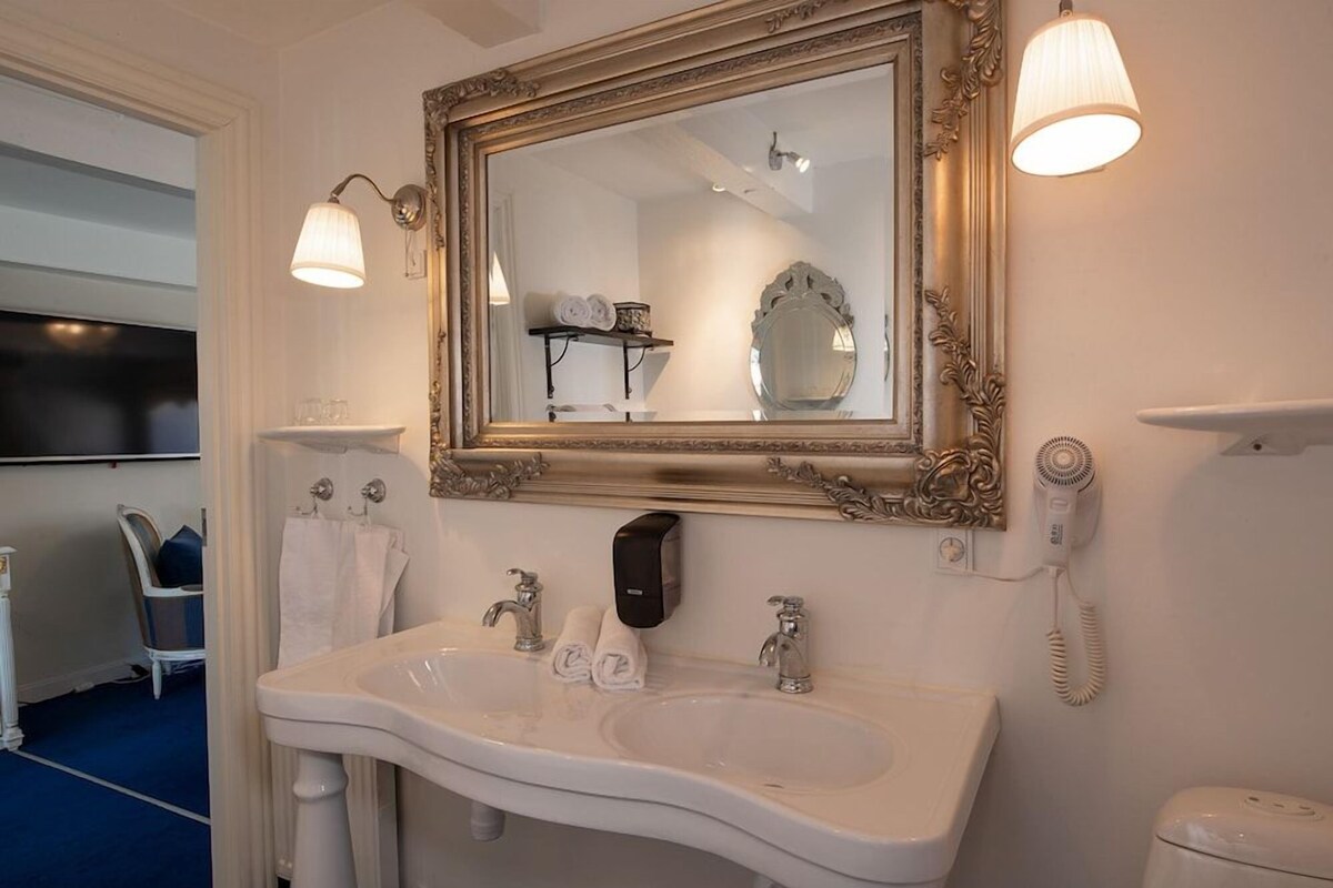 Un baño con un espejo grande y ornamentado, un lavabo blanco, dos dispensadores de toallas, un secador de pelo y un teléfono.