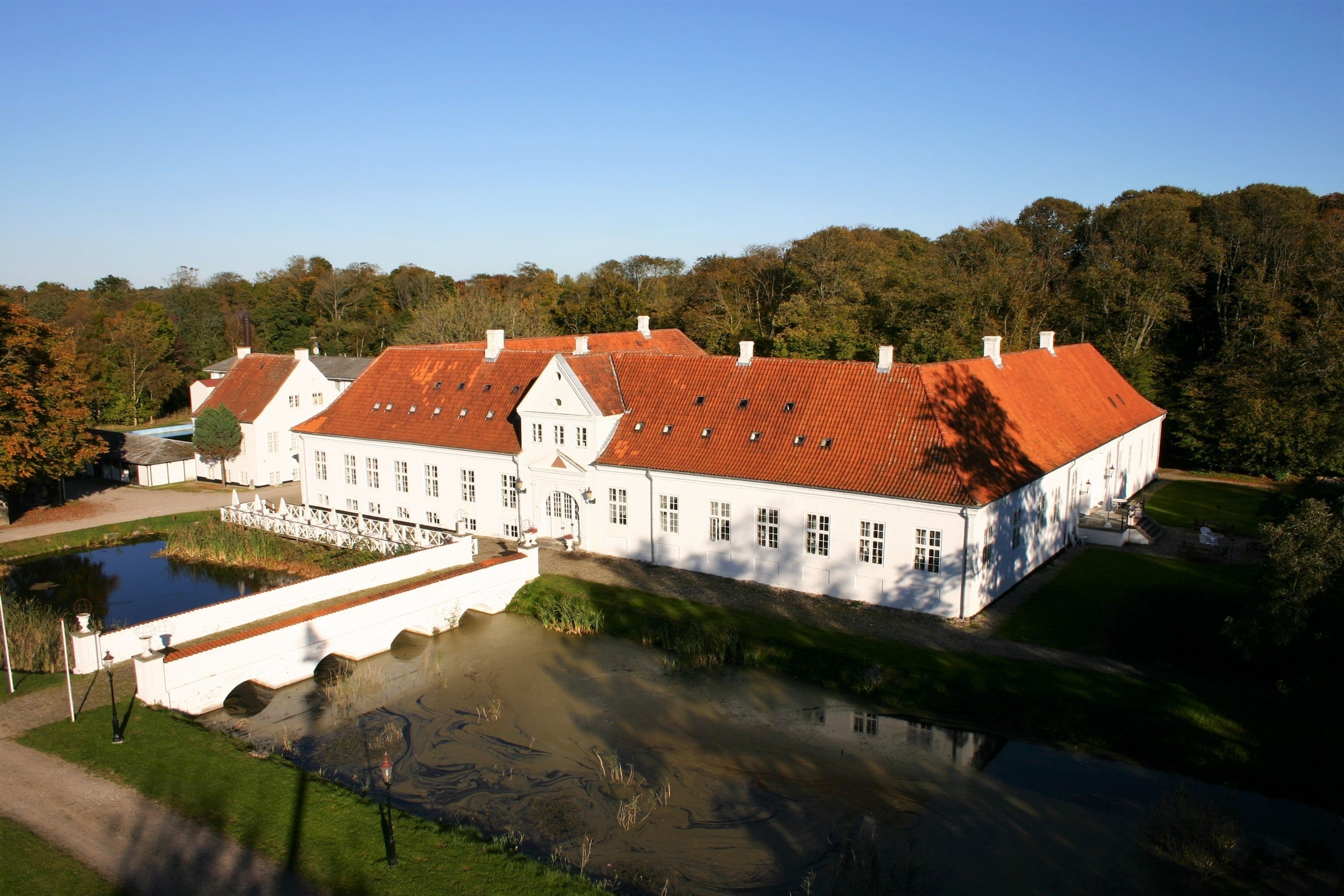 A large white building with a red roof, surrounded by a pond and trees.