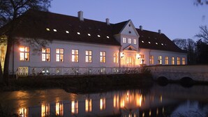 Un edificio amplio e iluminado con numerosas ventanas, un puente y una masa de agua tranquila que refleja las luces.