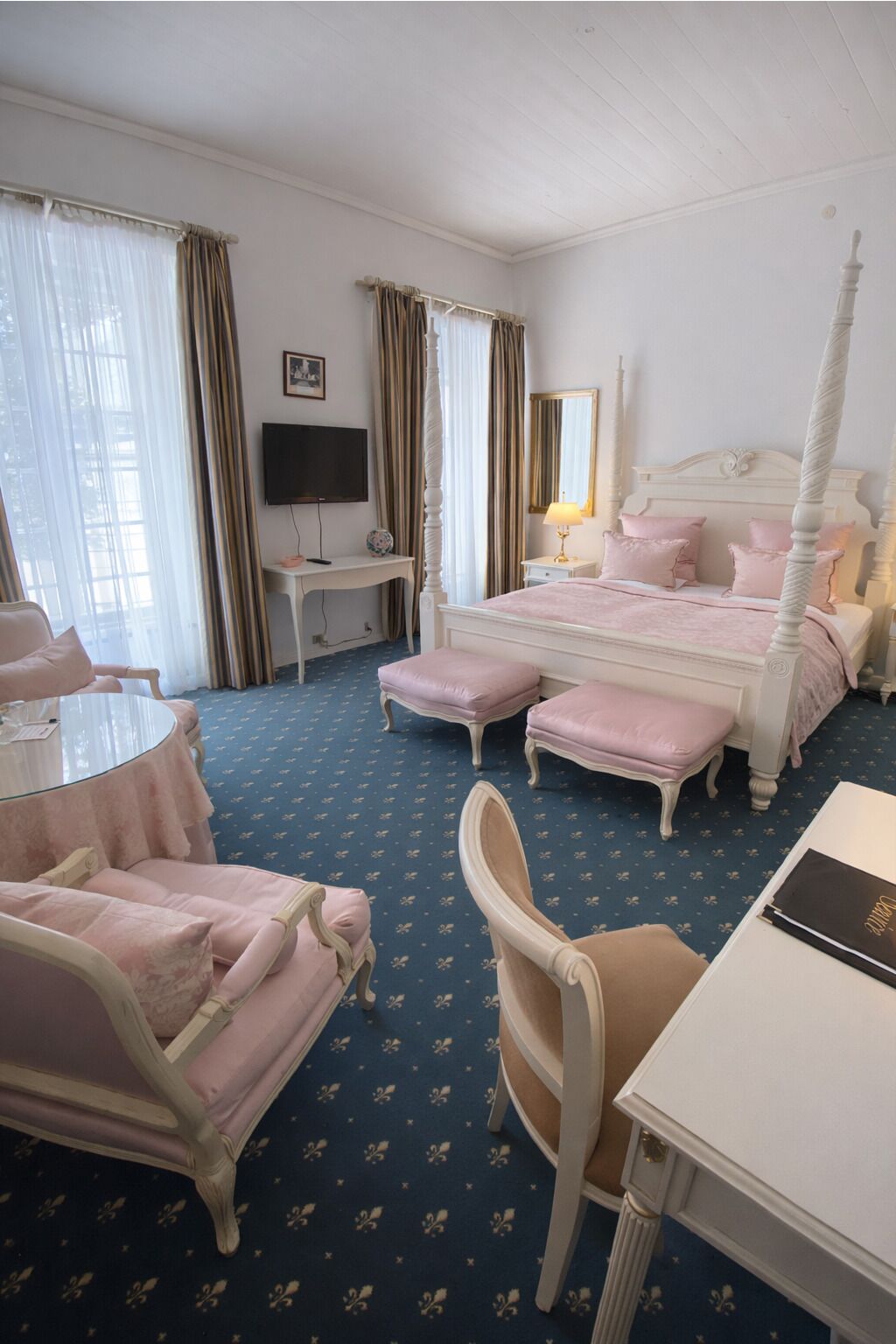 A hotel room with a four-poster bed, a desk with a lamp, a chair, and a television.