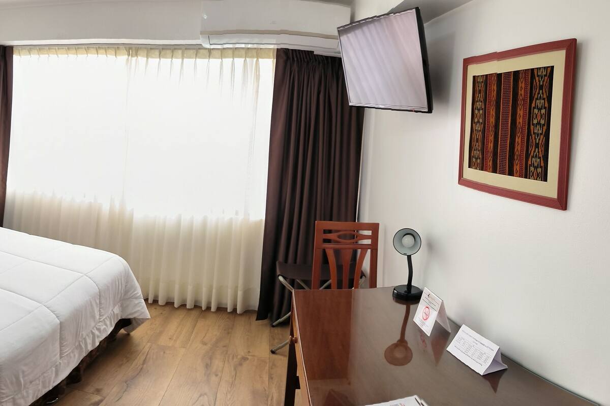A hotel room with a large bed, a bedside table with a phone, a desk with a mirror, and a TV mounted on the wall.