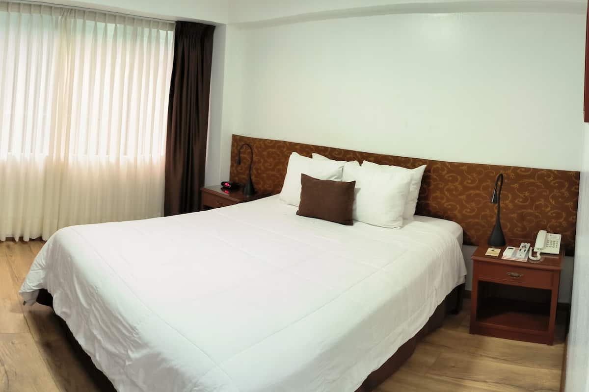 A hotel room with a large bed, a desk with paperwork, a chair, a nightstand with a phone, and a TV mounted on the wall.