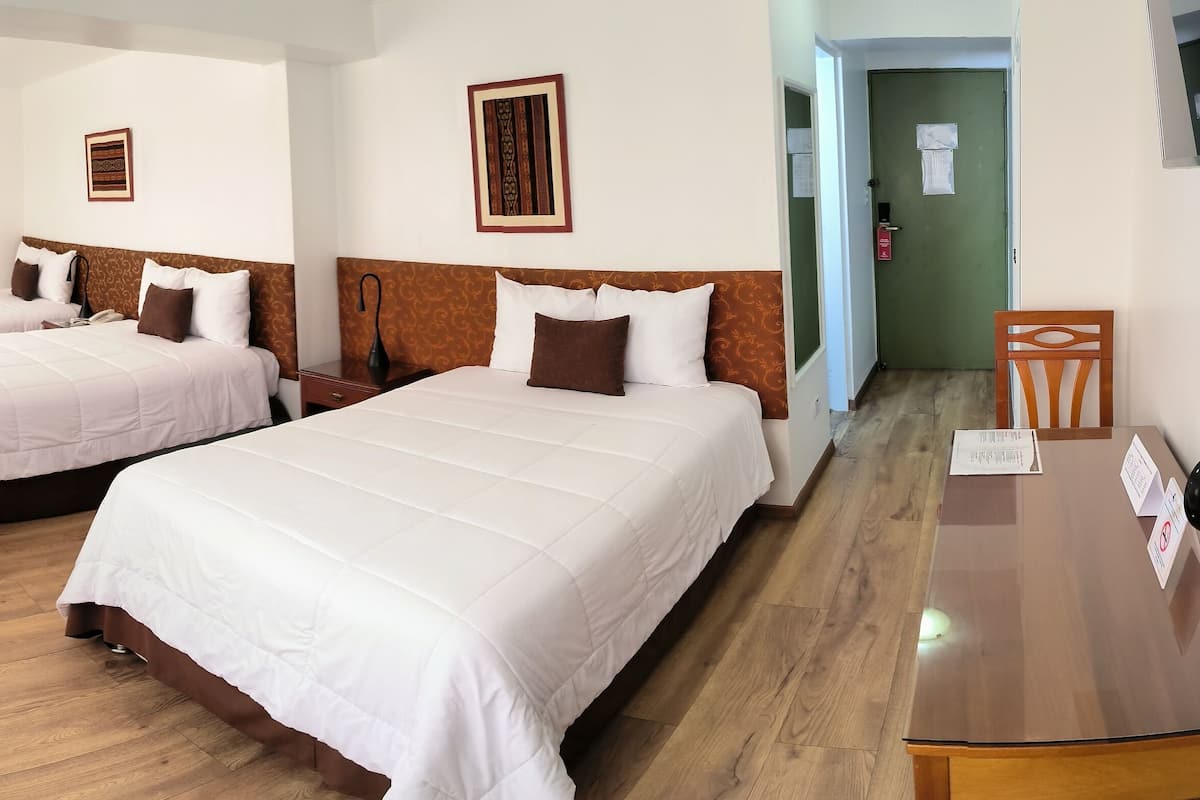 A hotel room with three beds, wooden flooring, and a desk with a chair.