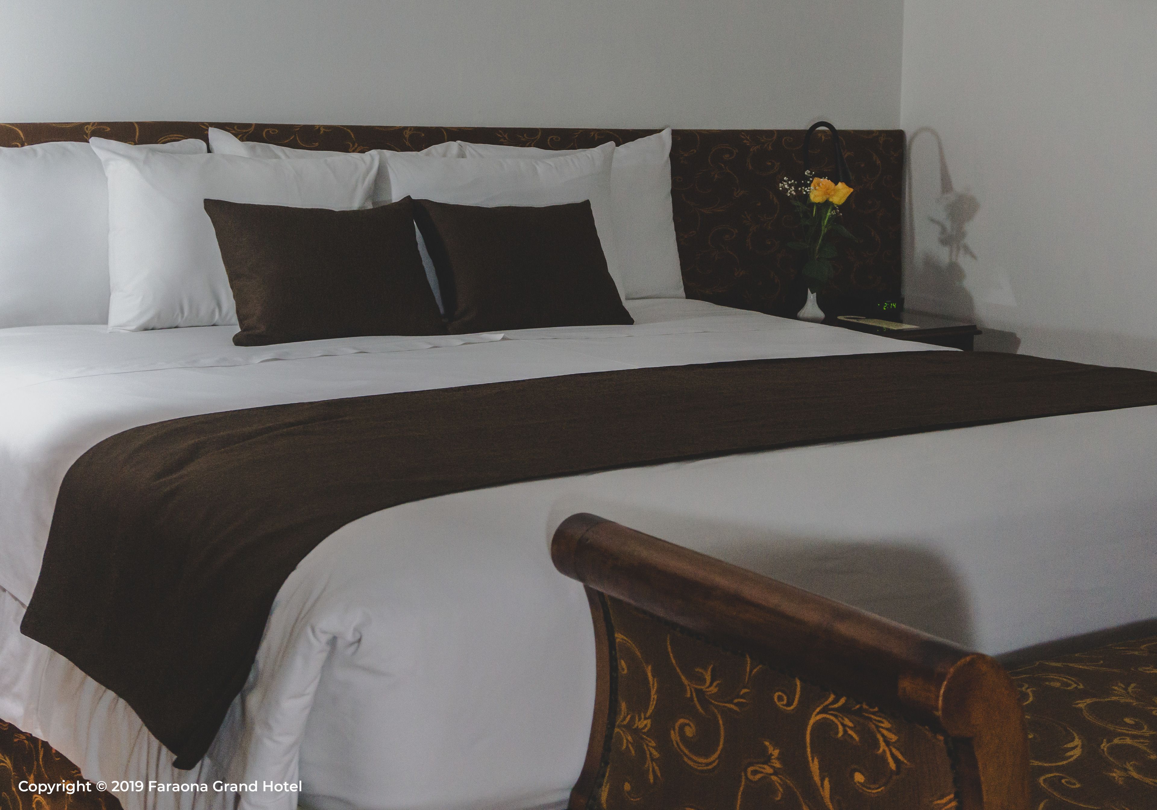 A neatly made bed with white and brown bedding, a wooden headboard with a patterned design, and a nightstand with a vase of flowers.