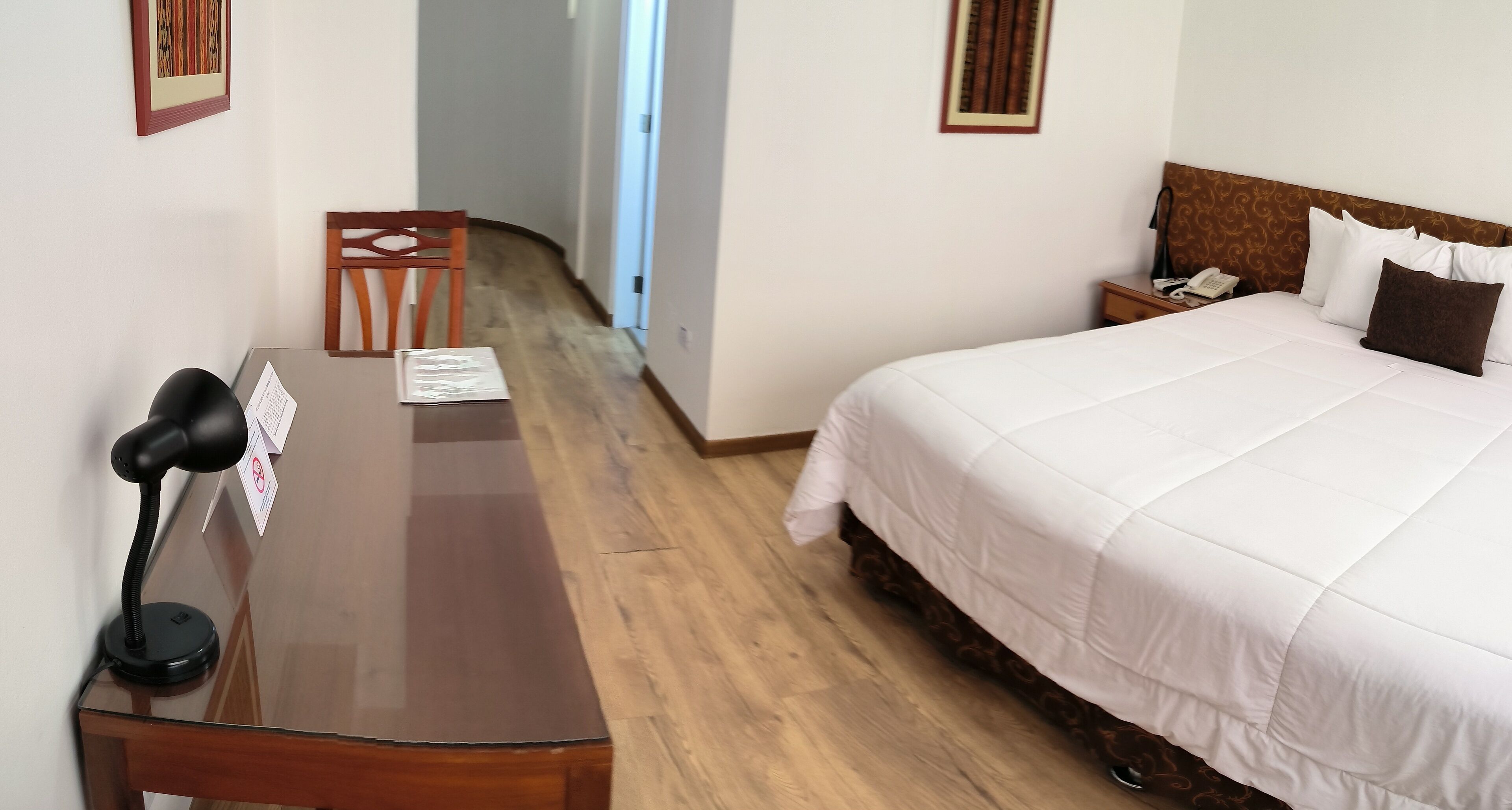 A hotel room with a wooden desk, a chair, a bed with white bedding, a nightstand with a phone, and a door leading to a hallway.