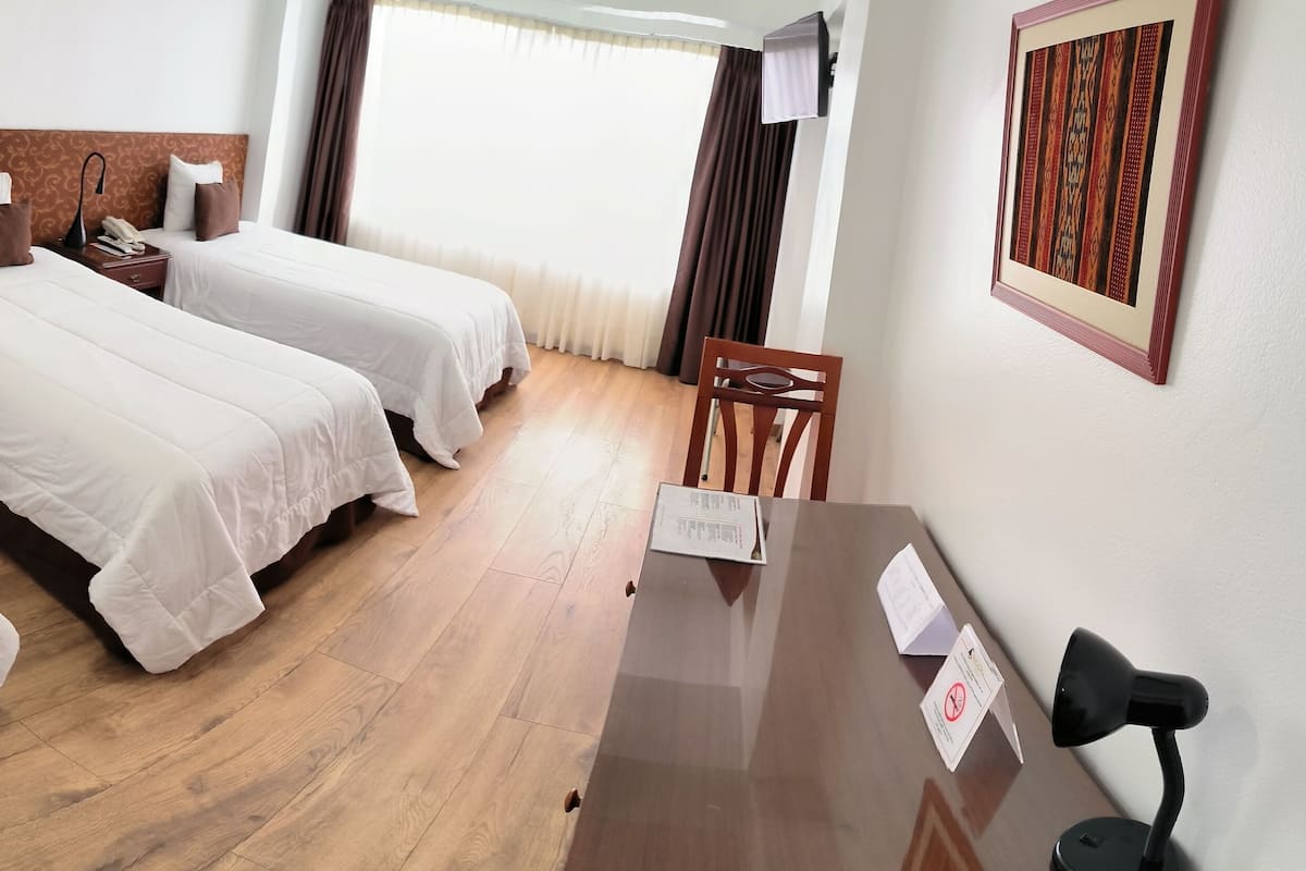 A hotel room with two single beds, a desk with a chair, a telephone, and a window with curtains.