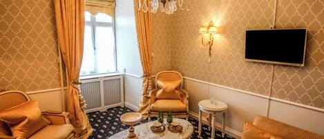 A room with a patterned carpet, a chandelier, a television mounted on the wall, and a seating area with a sofa and armchairs.