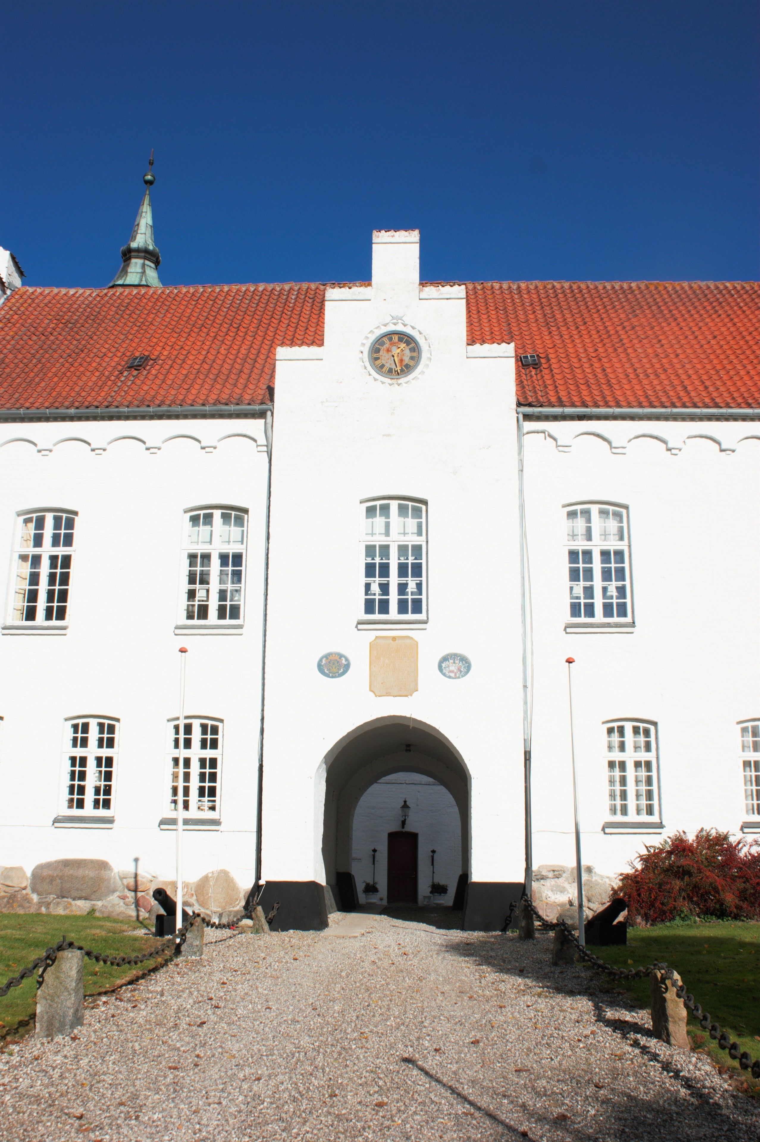 A white building with a red-tiled roof, a central archway, and a clock above it.