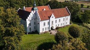 A large, white building with a red-tiled roof, surrounded by lush greenery and a clear sky.