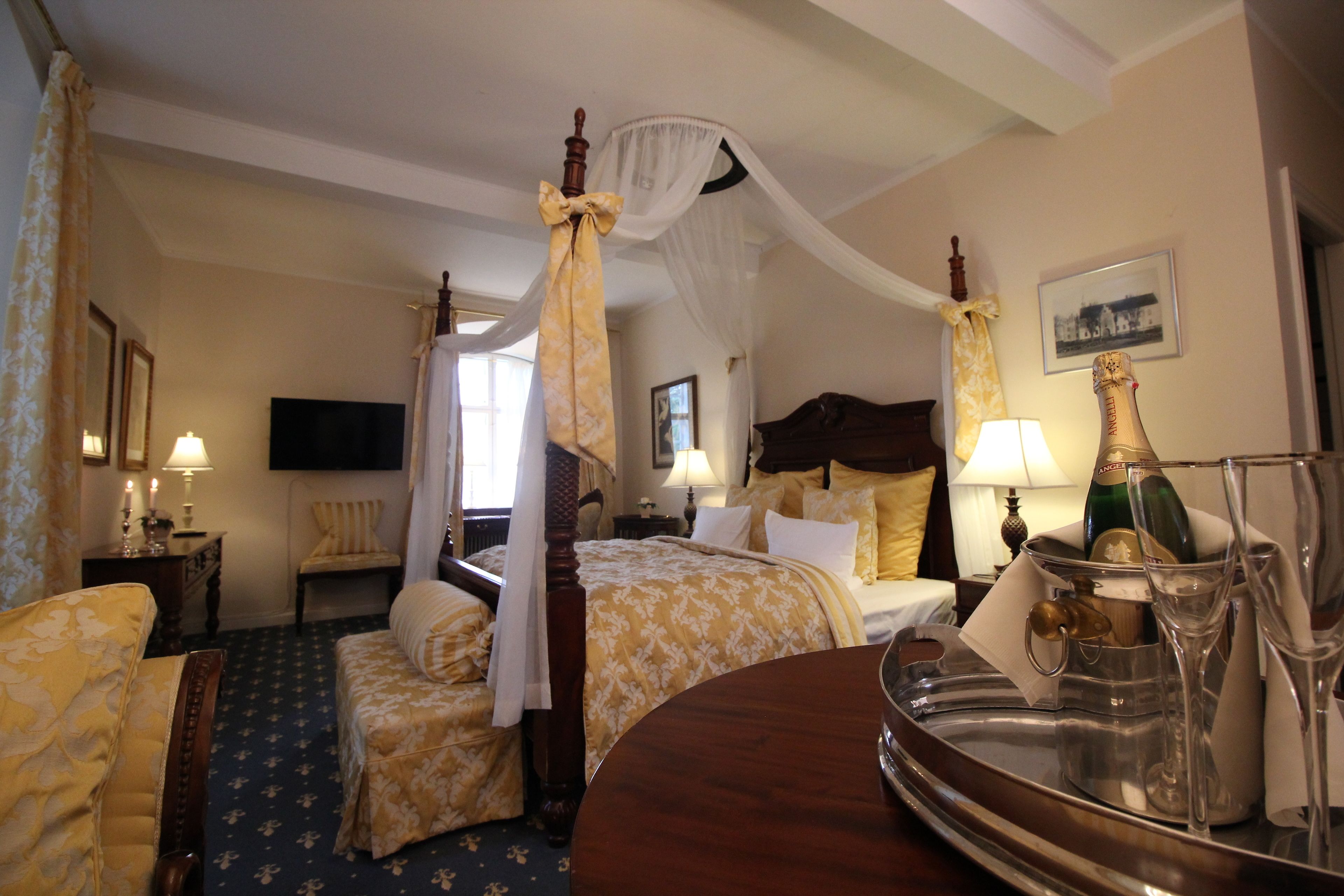 A hotel room with a four-poster bed, a canopy bed, a sitting area with a chair, a bedside table with a lamp, and a tray with champagne bottles and glasses.