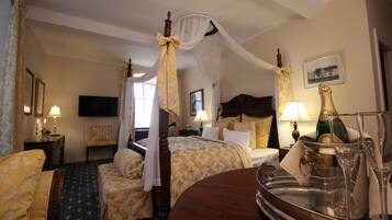 A hotel room with a four-poster bed, a canopy bed, a sitting area with a chair, a bedside table with a lamp, and a tray with champagne bottles and glasses.
