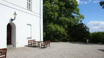 A white building with a gravel courtyard, wooden benches, and a large tree.