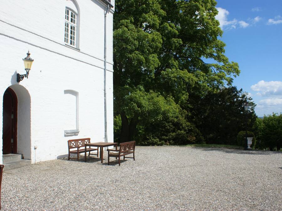 A white building with a gravel courtyard, wooden benches, and a large tree.