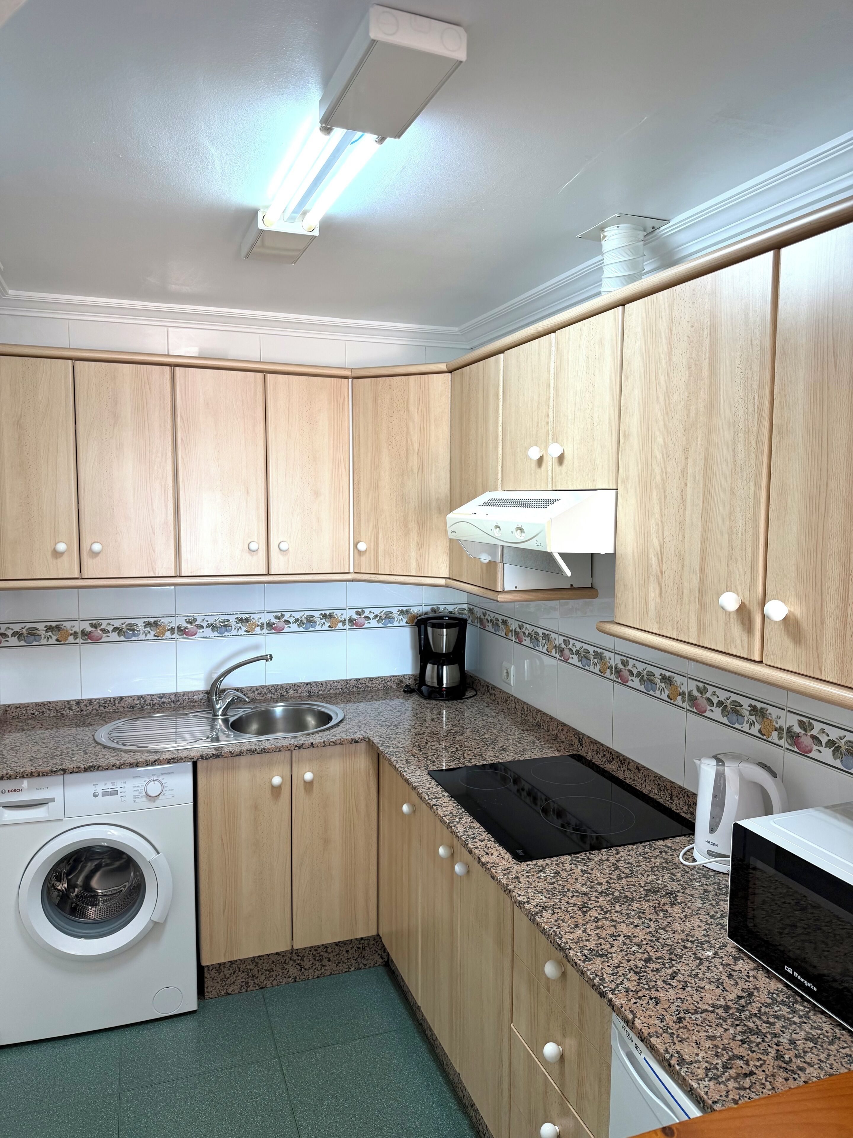 A compact kitchen with wooden cabinets, a built-in washing machine, a coffee maker, and a microwave.