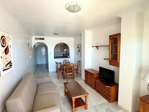 Sala de estar con sofá, una mesa de centro de madera, mueble para televisión y una zona de cocina con mesa y sillas.