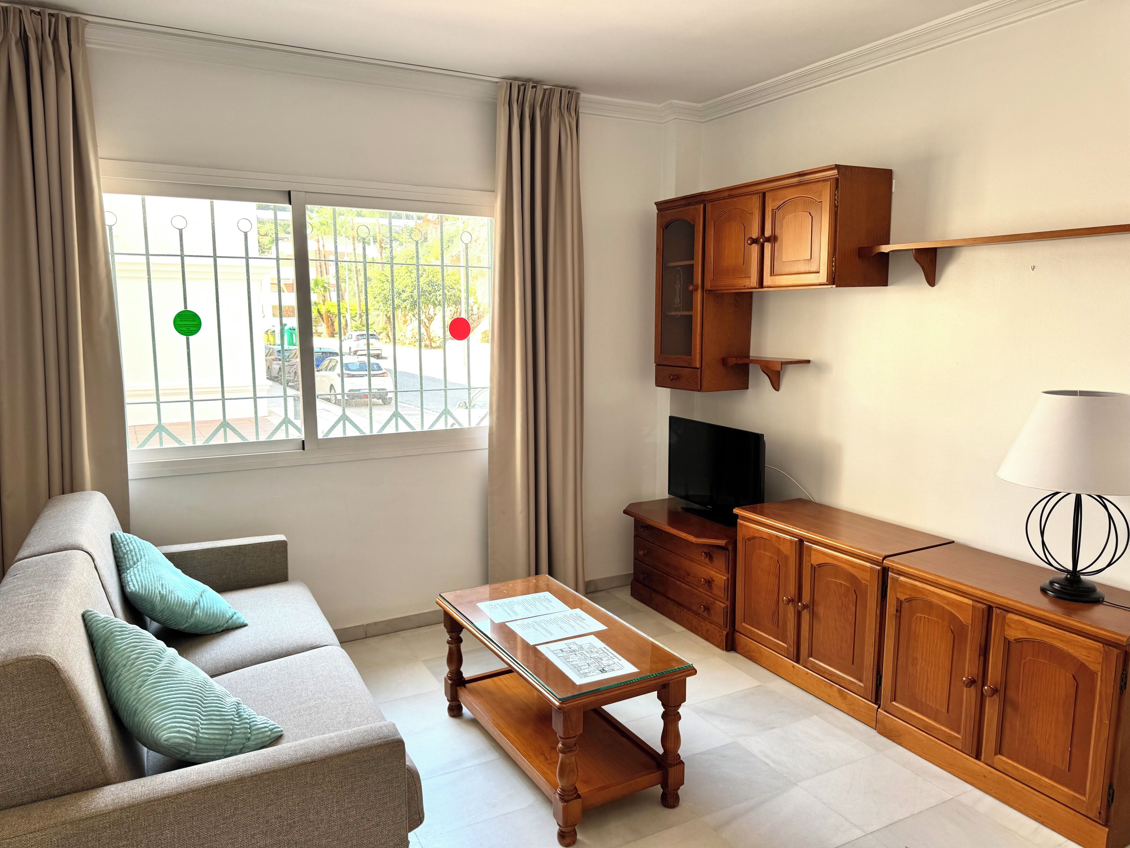 A living room with a sofa, a coffee table, a TV, wooden cabinets, and a window with a view of parked cars.