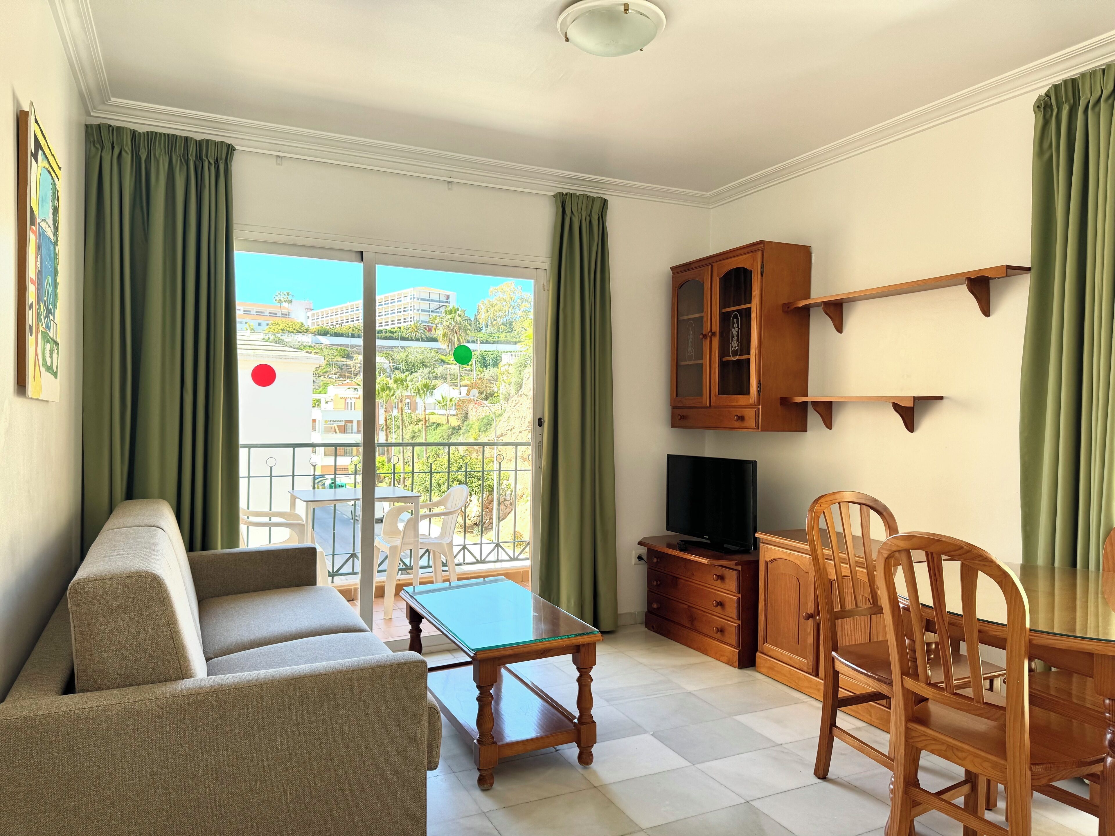 A living room with a sofa, a glass-top coffee table, a TV, wooden cabinets, and a balcony with curtains.