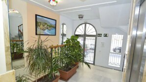 A hallway with potted plants, a painting on the wall, and a view of the outside through a glass door.