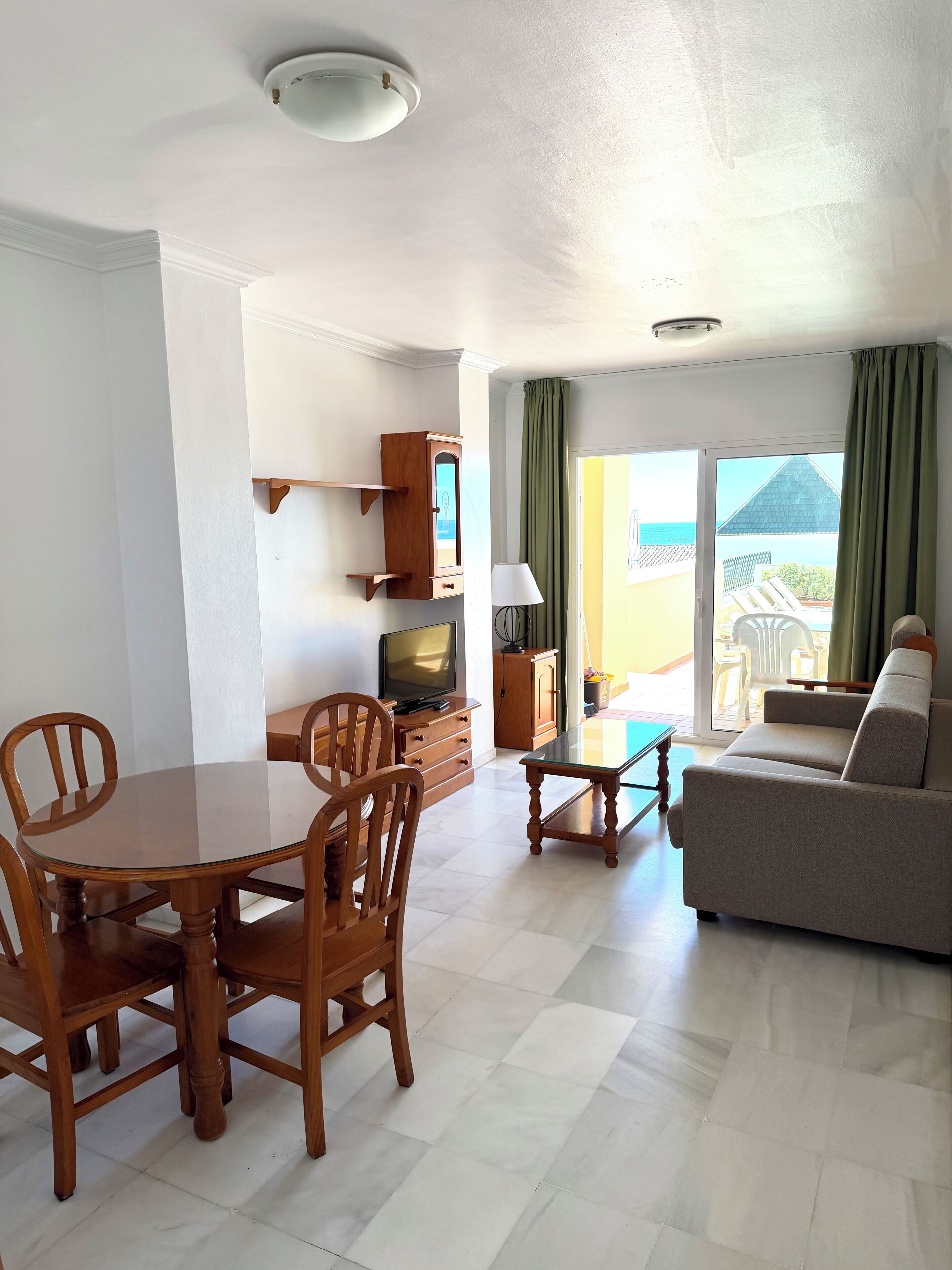 A living room with a dining table, chairs, a sofa, a TV, and a view of the sea through large windows.