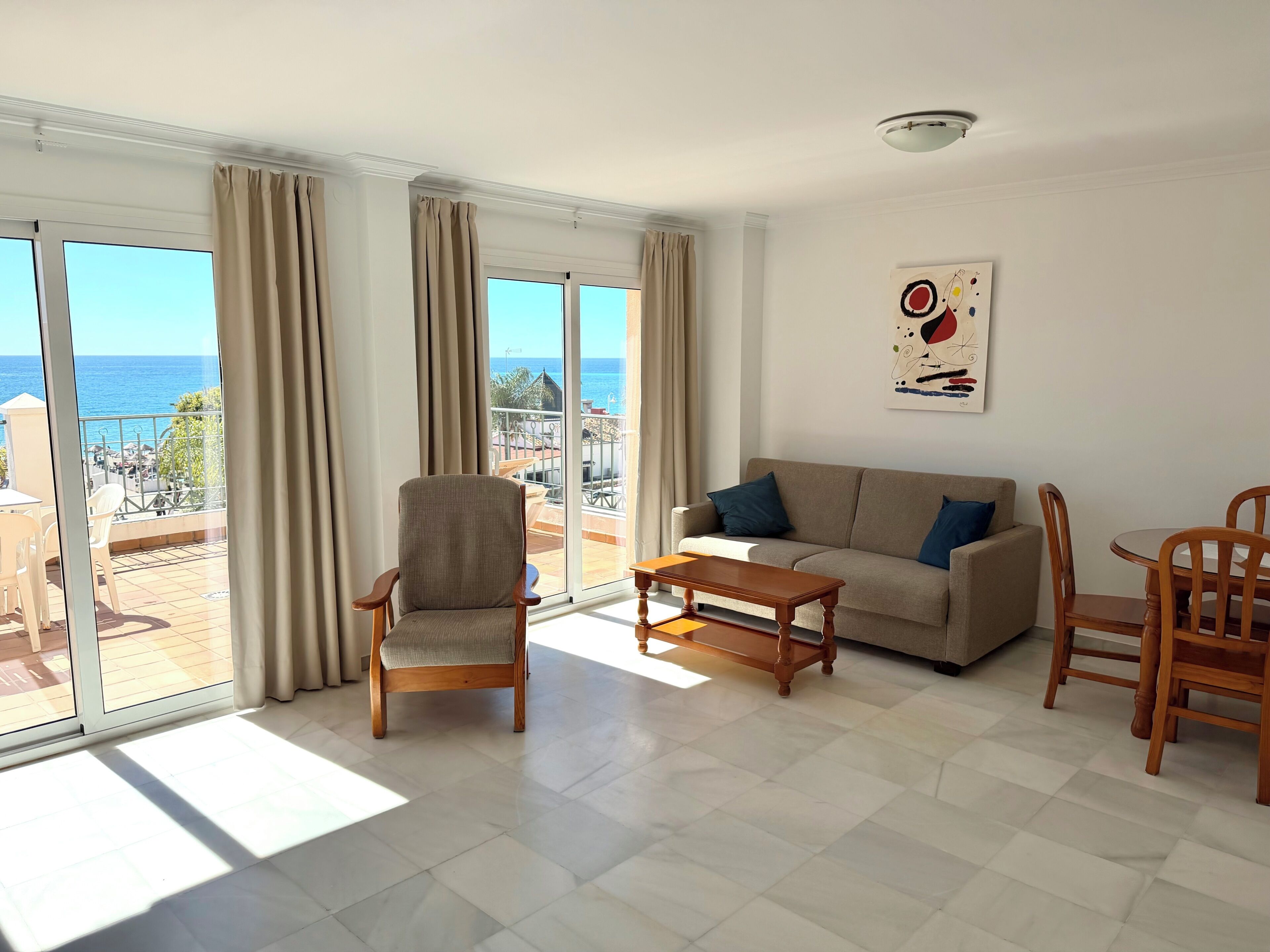 A living room with a sofa, armchairs, a coffee table, and a dining set. Large windows offer a view of the sea and a balcony.