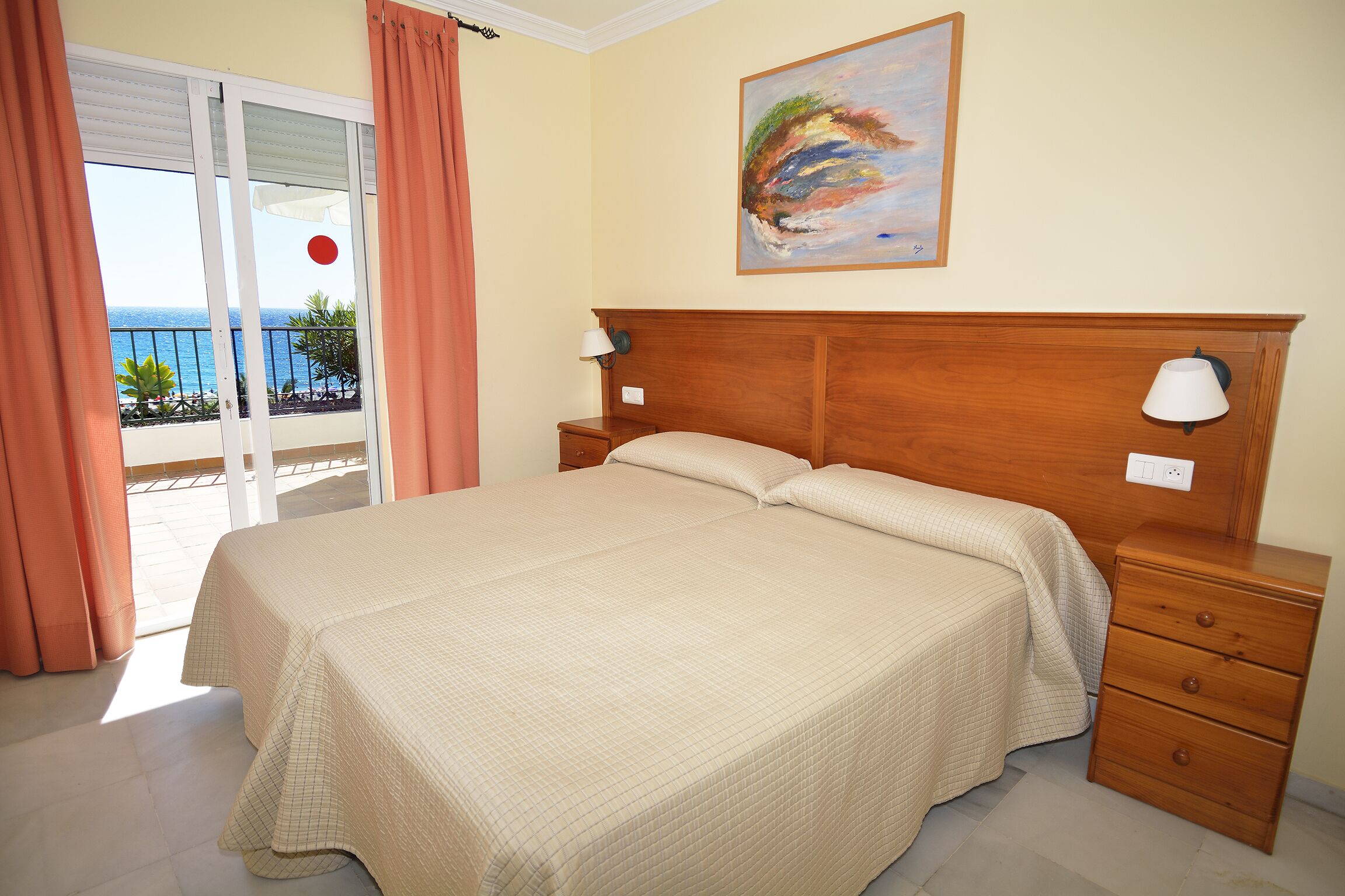 Ein Schlafzimmer mit Bett, Nachttisch, Lampe, einem Gemälde und Blick auf das Meer durch eine Schiebetür.