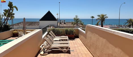 A rooftop terrace with lounge chairs, a small building, and a view of the sea.