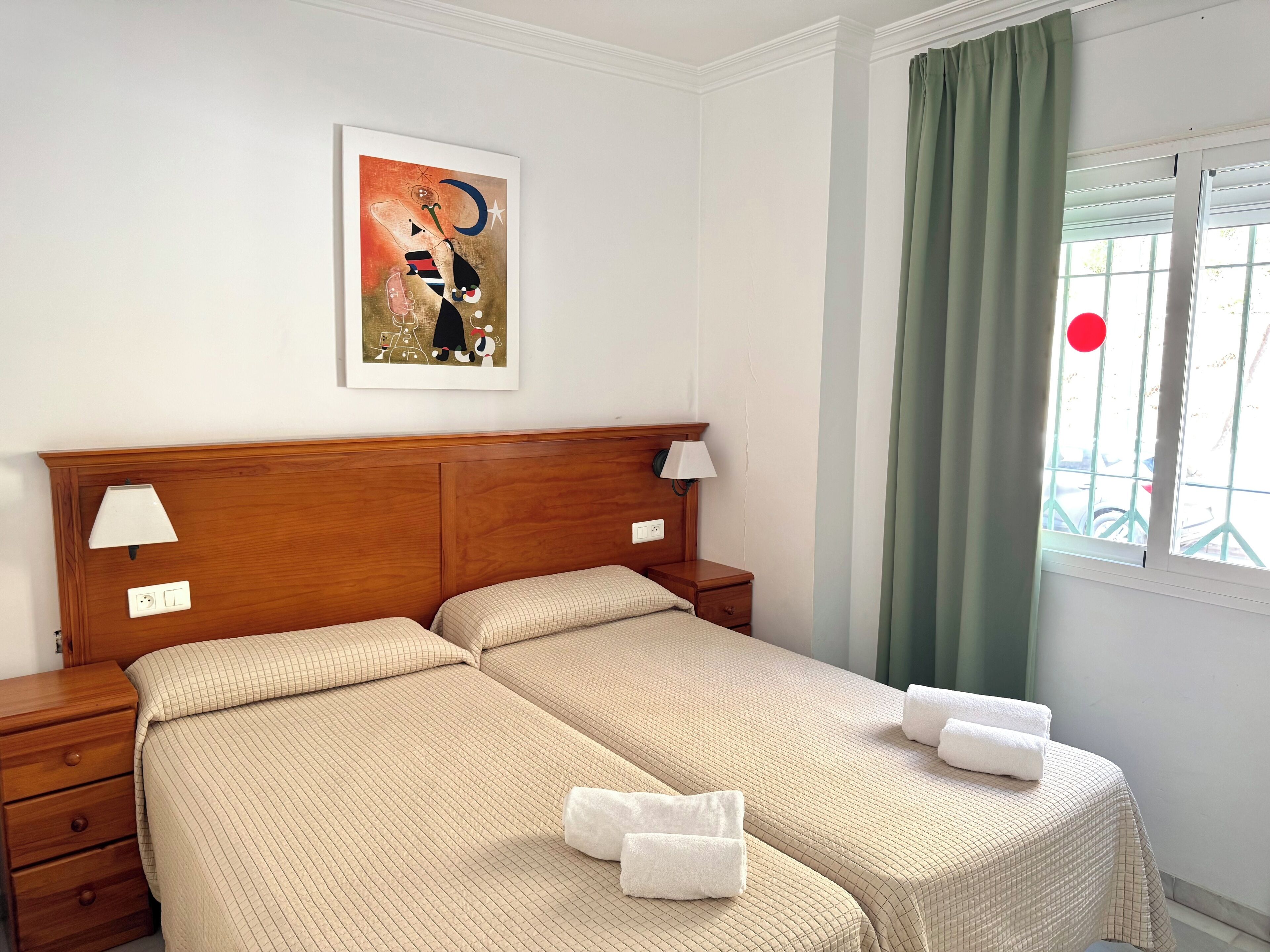 Uma cama de casal com cabeceira de madeira, duas mesas de cabeceira com abajures, um quadro pendurado na parede e uma janela com cortina verde.