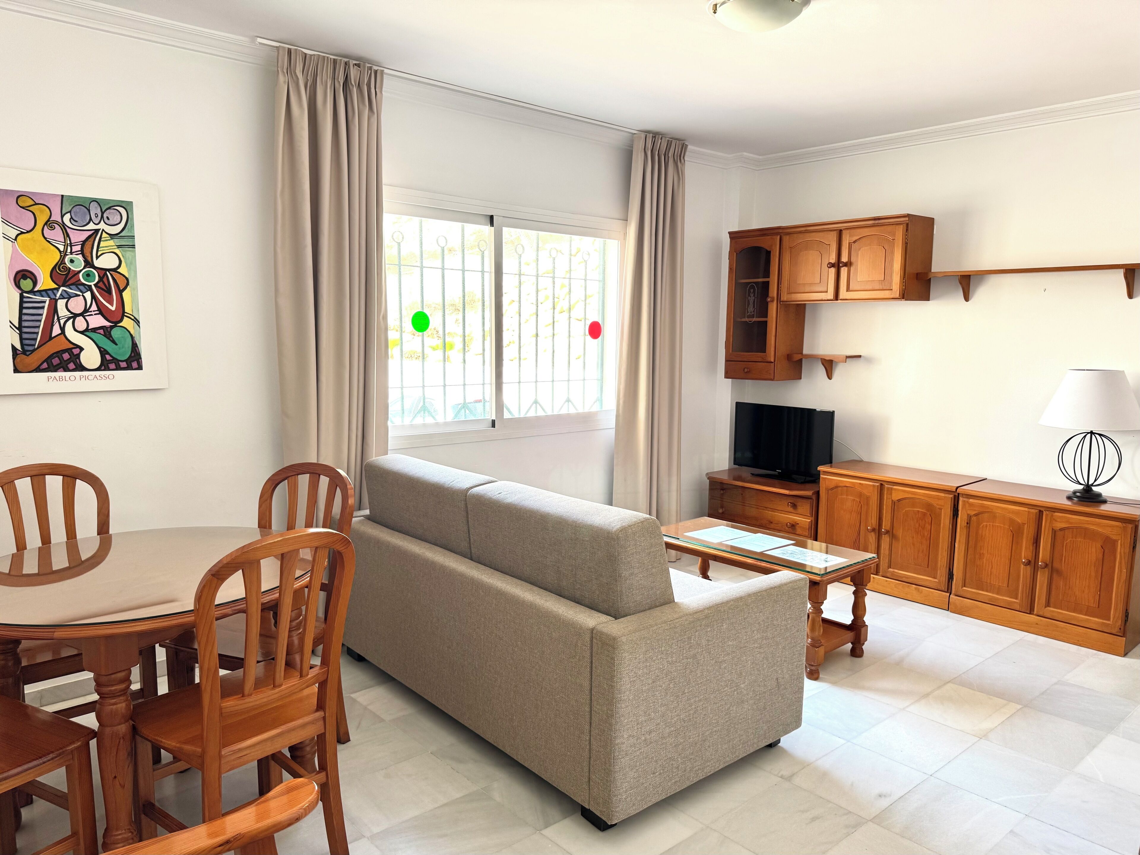 A living room with a sofa, dining table, chairs, a TV, wooden cabinets, and a window with curtains.