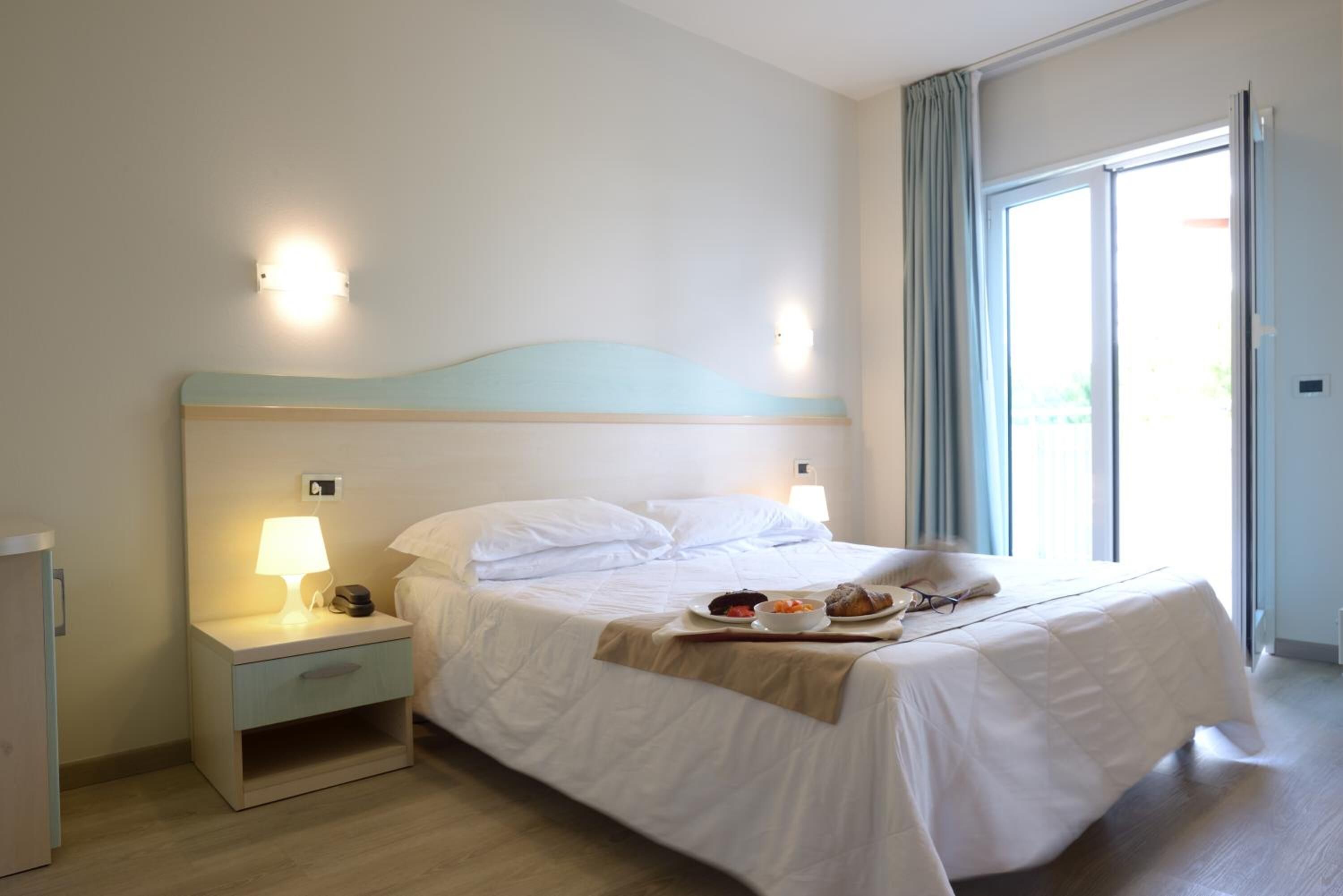 Ein Hotelzimmer mit Bett, Nachttischlampen, einem Tablett mit Frühstücksgegenständen und Blick ins Freie.