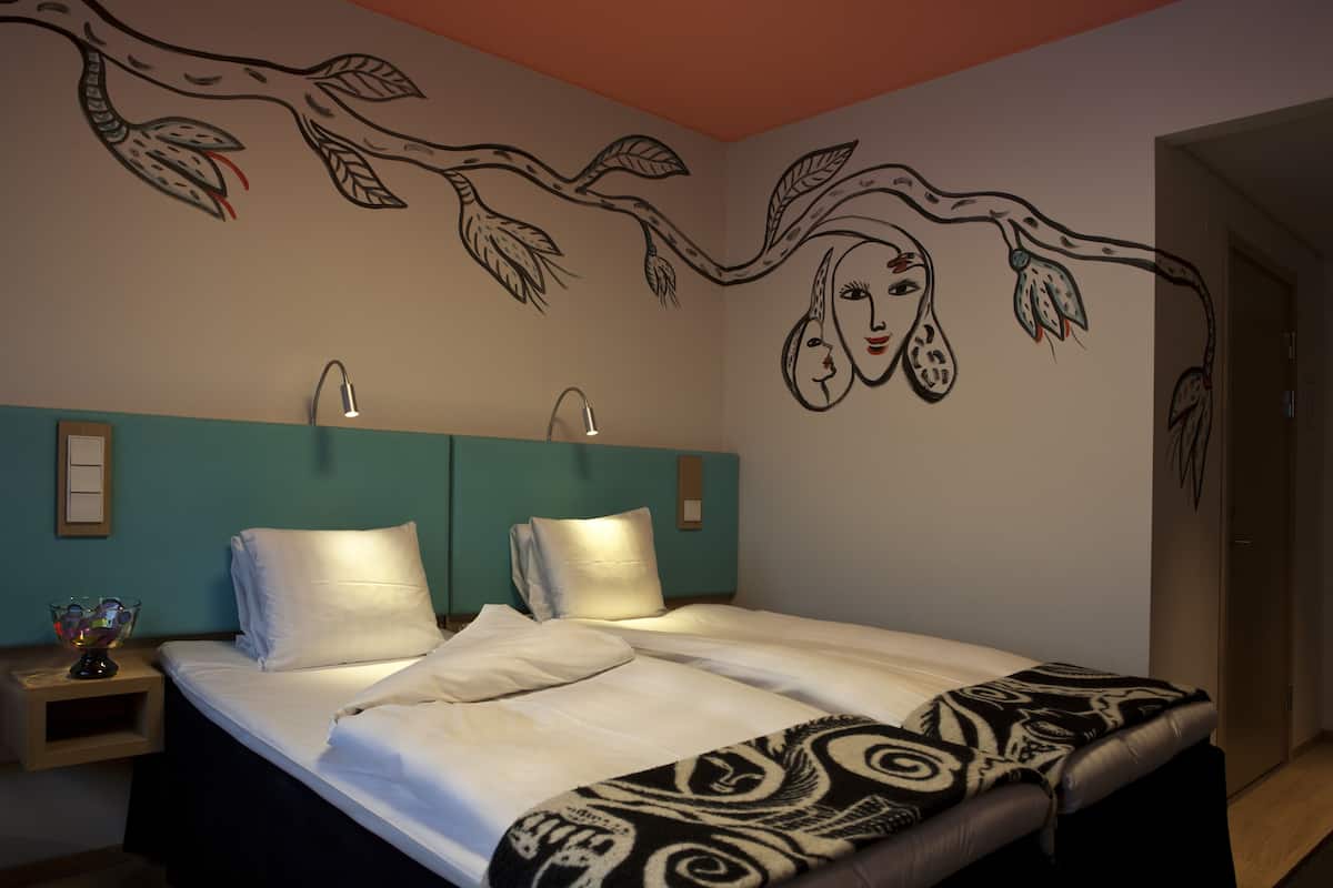 Un dormitorio con cama, dos lámparas de noche, un mural en la pared y una mesita de noche con un cuenco de vidrio.
