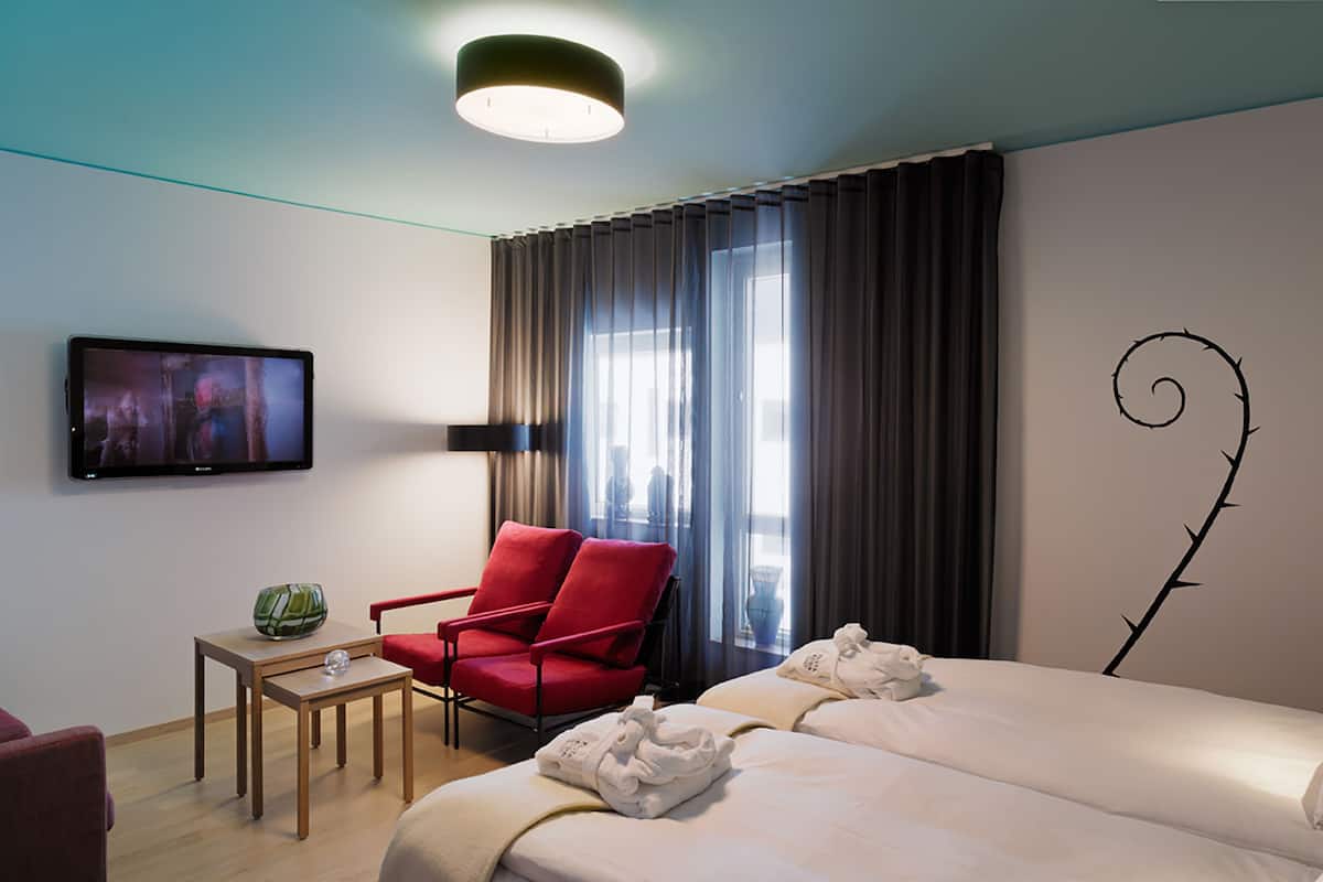 Una habitación de hotel con una cama, un sillón rojo, una mesita con un objeto verde, un televisor en la pared y una ventana con cortinas.