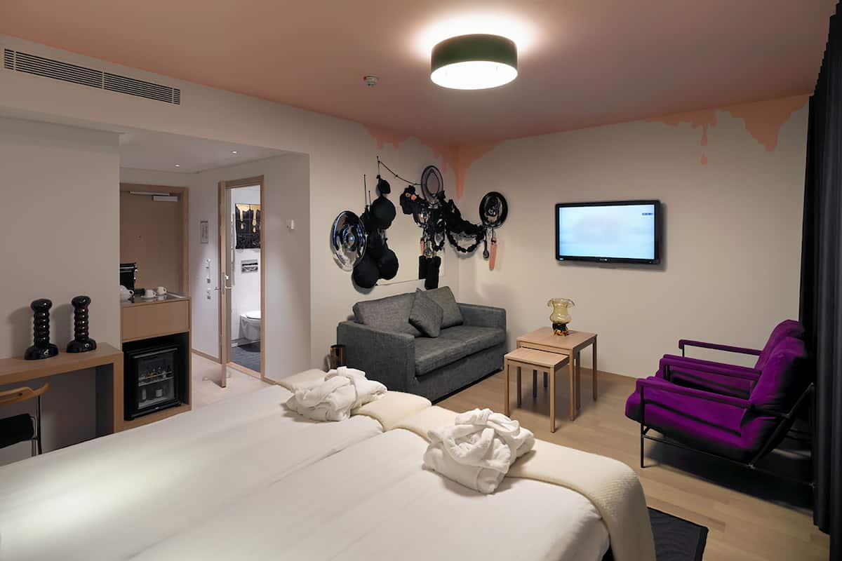 Una habitación de hotel moderna con una cama grande, un sofá gris, una pequeña mesa, un sillón morado y un televisor en la pared.