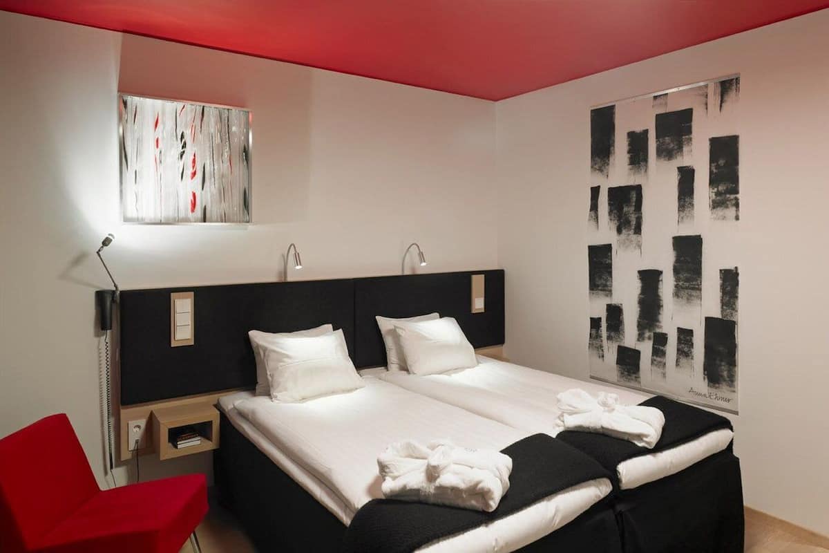 Una habitación de hotel moderna con una cama grande, una silla roja y arte abstracto en la pared.