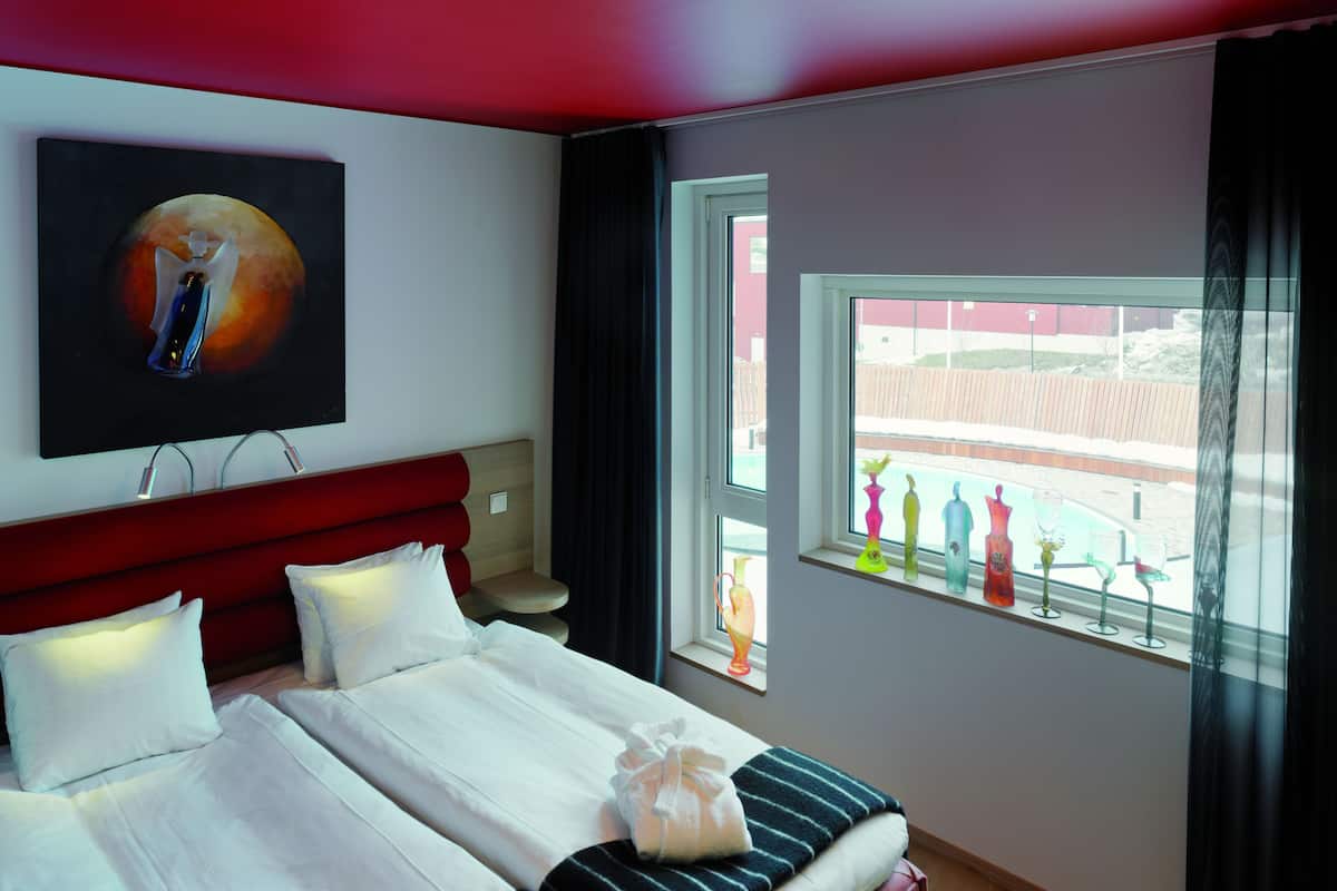 Habitación de hotel con cama, cabecera, cuadro, ventana con vistas y mesita de noche.