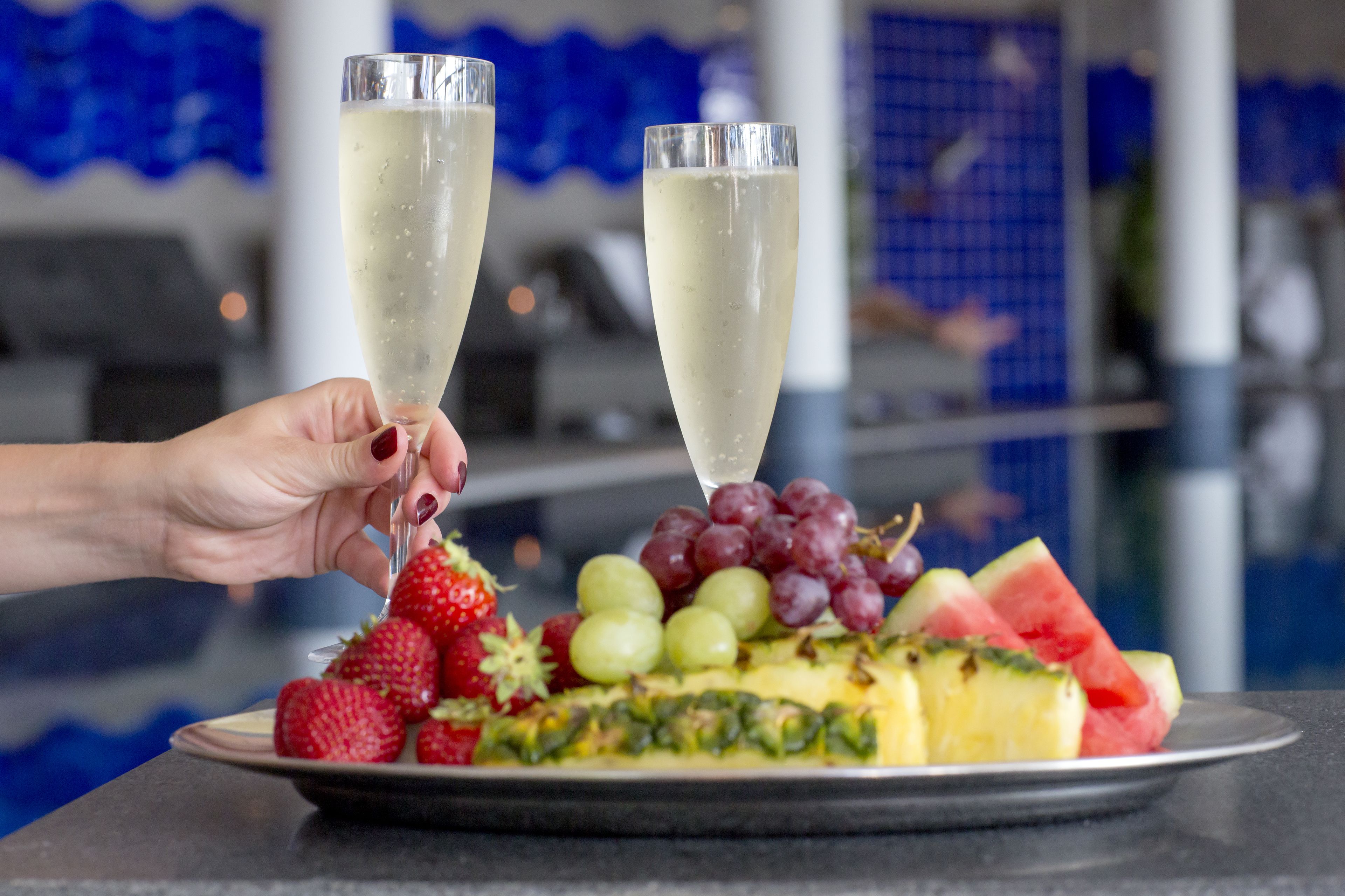 Een hand die twee champagneglazen vasthoudt boven een fruitschaal met aardbeien, druiven en ananas.