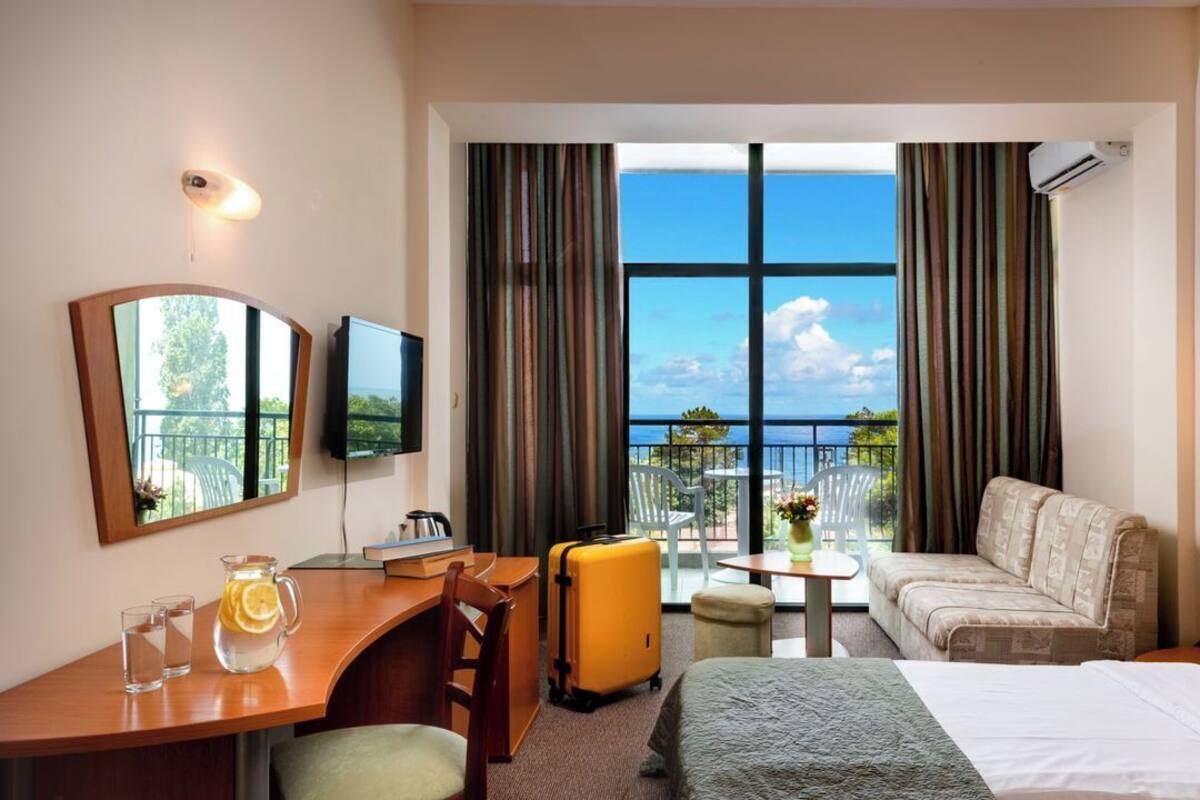 A hotel room with a bed, a desk, a chair, a sofa, a TV, and a window with a view of the sea.