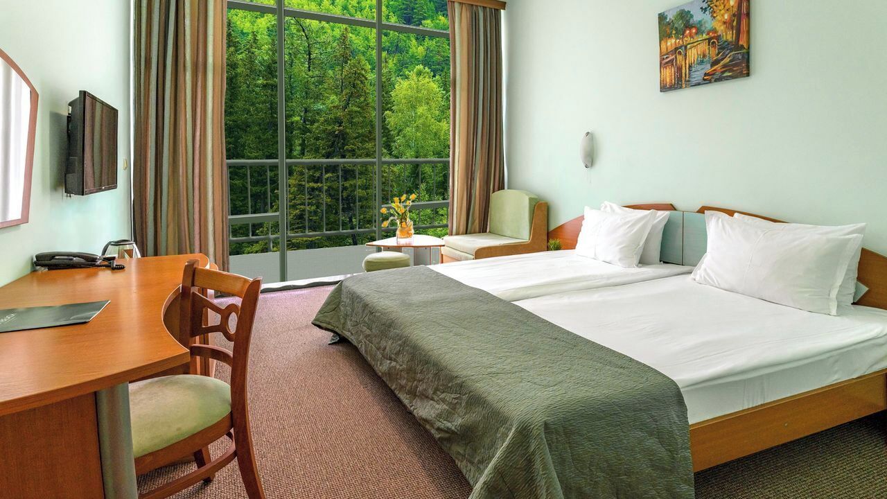 Una habitación de hotel con un ventanal grande, un escritorio, dos camas y un cuadro en la pared.