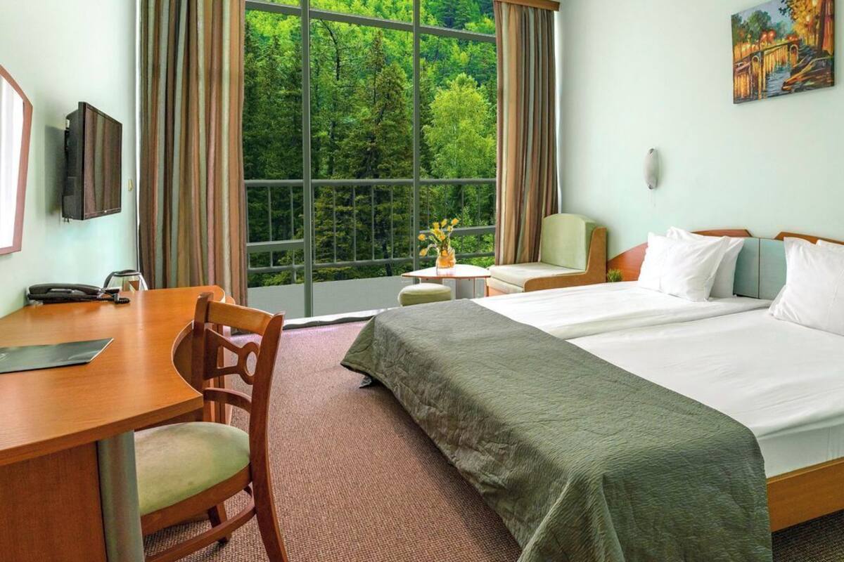 A hotel room with a large bed, a desk with a chair, a small table with a vase, and a painting on the wall.