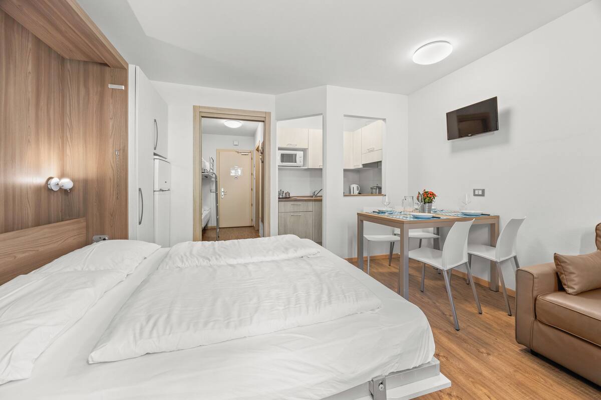 A modern hotel room with a large bed, a desk with chairs, a kitchenette, and a TV mounted on the wall.