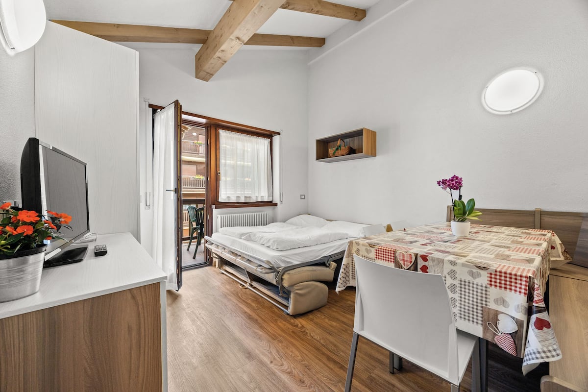 A compact room with a bed, a small table with a tablecloth, a chair, a desk with a computer and a plant, and a window with a view of a balcony.