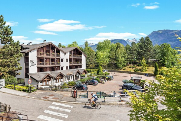 A hotel with a ski lift, a parking area, and a cyclist on the road. - Aparthotel Des Alpes (Cavalese)
