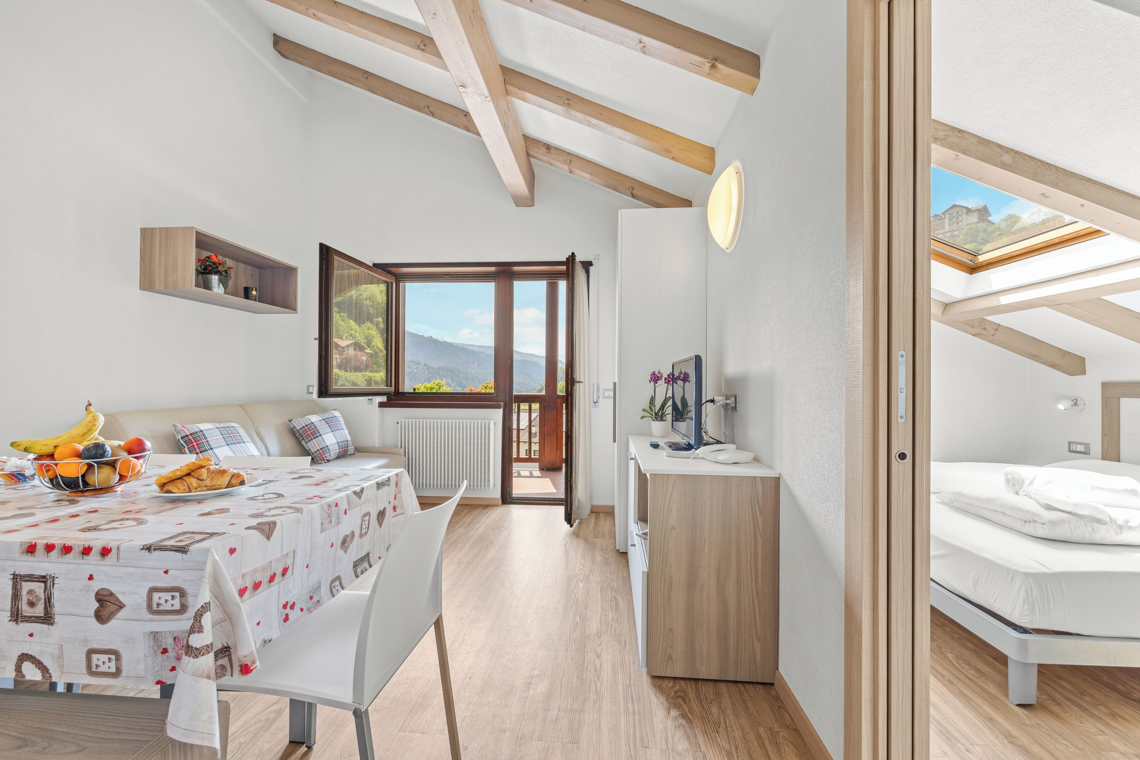 Ein modernes, helles Zimmer mit Holzbalkendecke, einem Essbereich mit Tisch für zwei Personen, einem Bett mit Oberlicht und Blick auf Grünfläche.