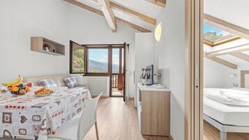 Ein modernes, helles Zimmer mit Holzbalkendecke, einem Essbereich mit Tisch für zwei Personen, einem Bett mit Oberlicht und Blick auf Grünfläche.