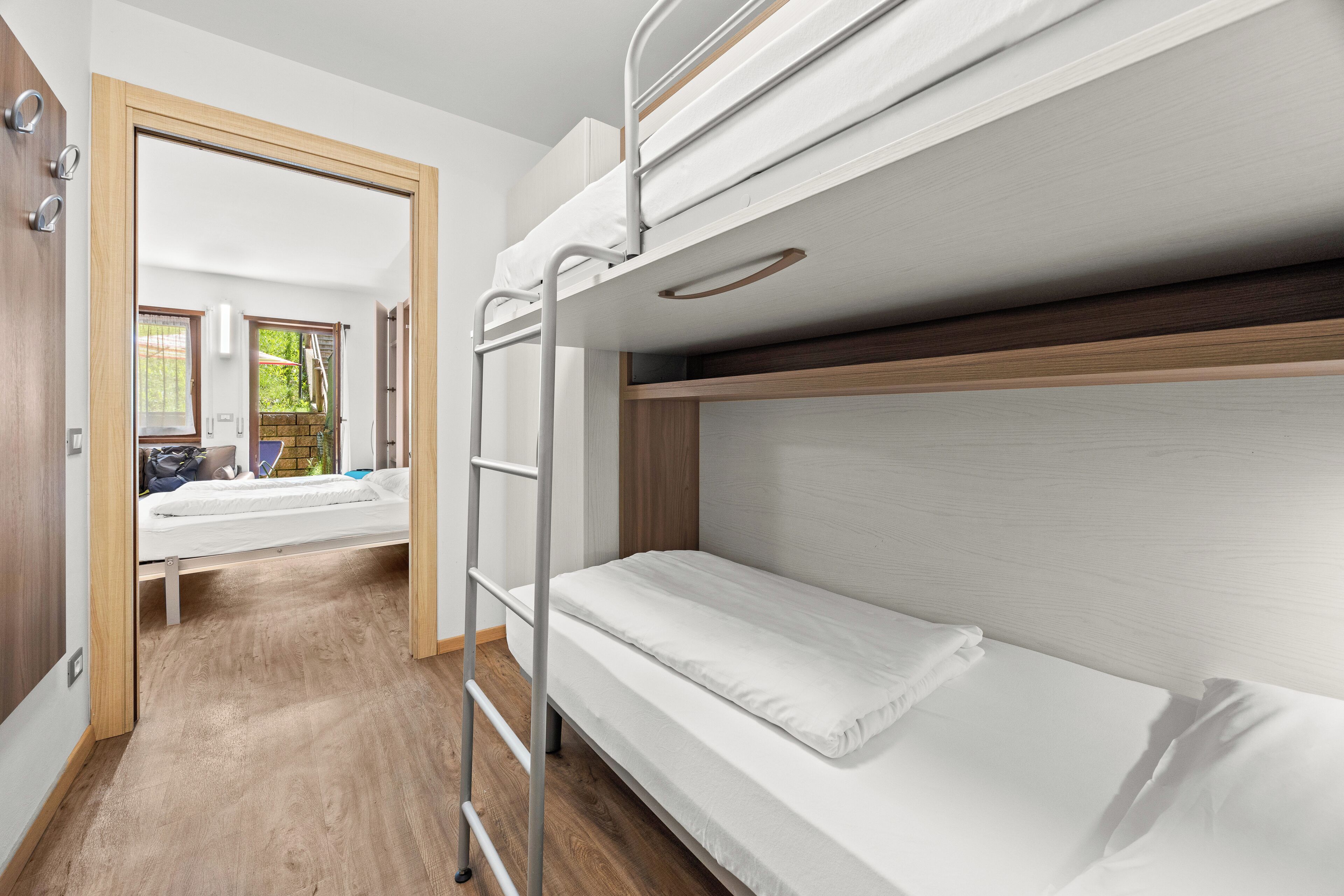 A bunk bed room with a wooden floor, a ladder, and a view of a garden through an open door.