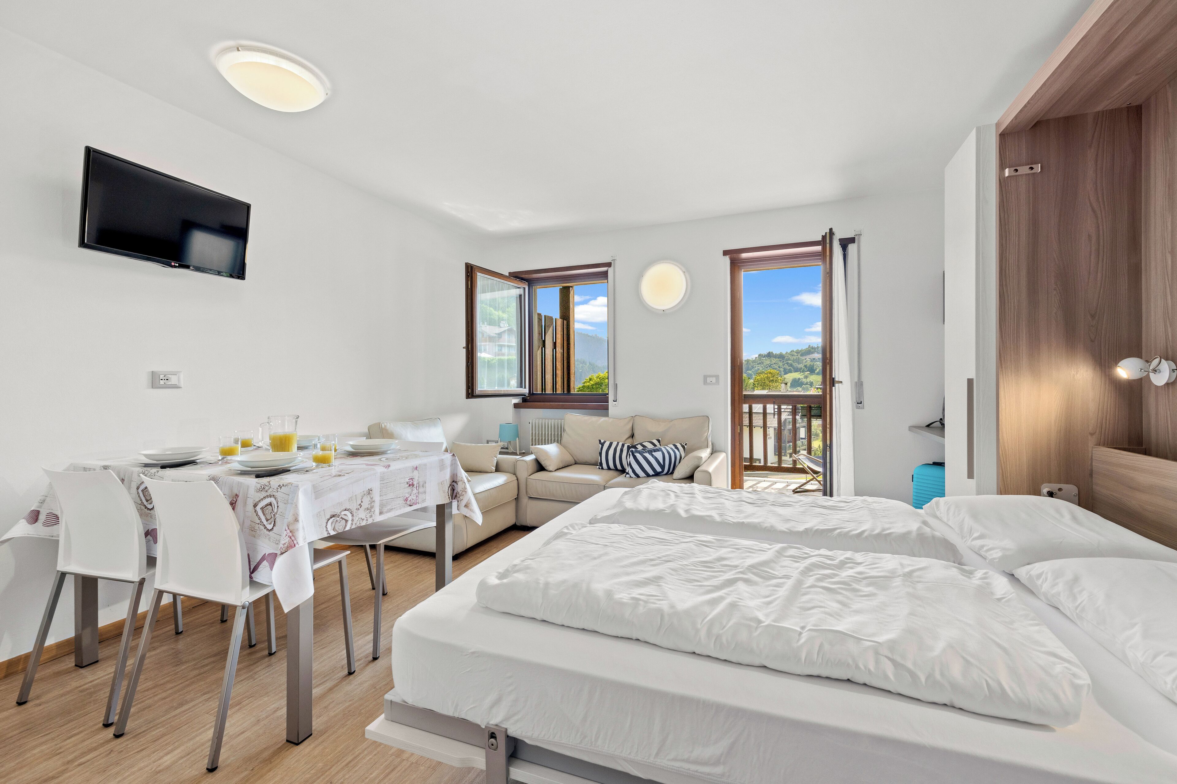 A modern room with a bed, a dining table with chairs, a TV mounted on the wall, and large windows offering a view of the outdoors.